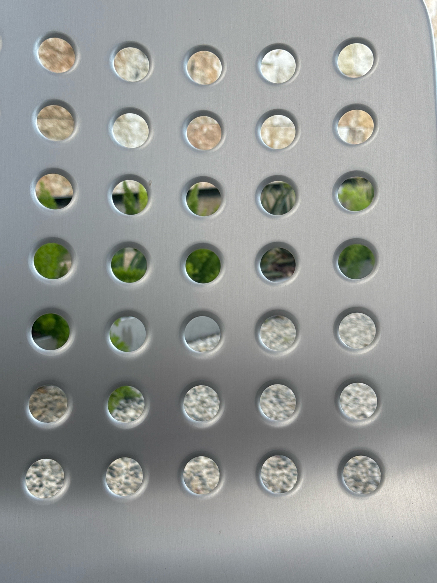 The image shows a close-up of a gray perforated surface, possibly a chair or a screen, against a blurred background. The perforations are circular and arranged in a grid pattern. Through the holes, glimpses of an outdoor environment are visible, including textured stone or brickwork, green foliage, and gravel. The lighting suggests it is daytime. The location is specified as Strovolos, Cyprus. There are no people or discernible actions in the image.