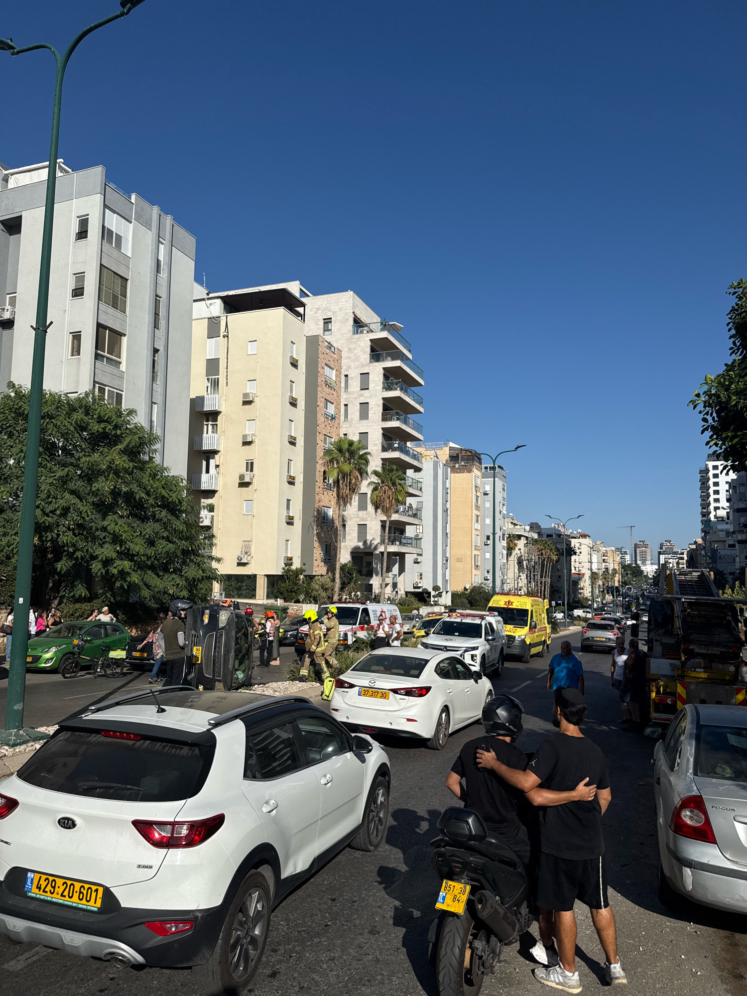 In the city of Netanya, Israel, on a clear, sunny day, a street scene unfolds with the aftermath of an apparent traffic accident. A black car is overturned on its side in the middle of the road, surrounded by emergency personnel in yellow and orange uniforms, likely firefighters and paramedics. Several other vehicles, including a white SUV, a green car, and a yellow ambulance, are present, suggesting a significant disruption. People are gathered on the sidewalk and near the accident site, observing the scene. The environment is urban, with multi-story buildings lining the street, some with balconies and air conditioning units. Palm trees are visible, indicating a warm climate. The sky is a vivid blue. In the foreground, two men stand with their arms around each other, one on a motorcycle and the other standing next to him, appearing to be offering comfort. The overall impression is one of a chaotic event that has temporarily halted the flow of traffic and drawn the attention of onlookers.