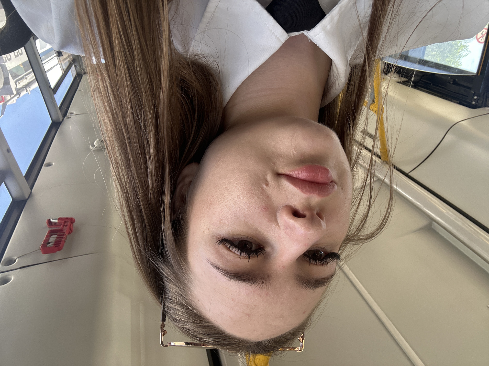 A young woman with long, light brown hair is taking a selfie. She is wearing a white collared shirt and a black tie. Her head is tilted slightly, and she is looking at the camera. She has long, dark eyelashes. Behind her, the interior of a vehicle is visible, including a yellow pole and part of a window. The ceiling of the vehicle is white and appears to have lights and a red plastic fixture. The lighting suggests it is daytime.