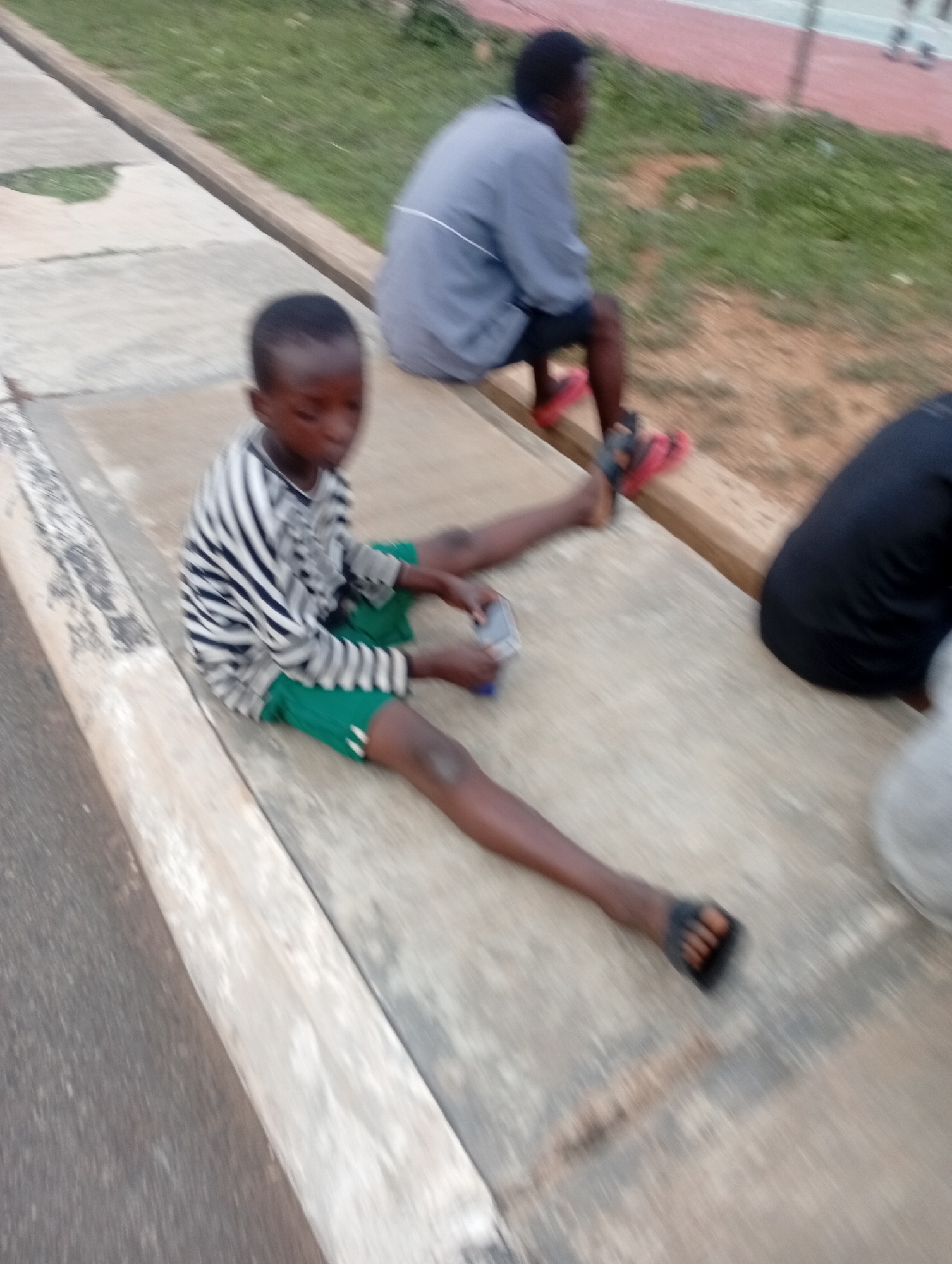 In the city of Jos, Nigeria, a young Black boy is sitting on a concrete ledge. He is wearing a black and white striped long-sleeved shirt and green shorts. His left hand is holding a small, light-colored object. To his right, another Black person in a grey shirt and dark shorts is sitting on the same ledge, with red flip-flops on their feet. Further to the right, a third person, whose body is mostly out of frame, is wearing a black shirt and is partially visible. The background consists of green grass and what appears to be a red track or court area in the distance. The scene is outdoors, likely in a public space like a park or sports facility.