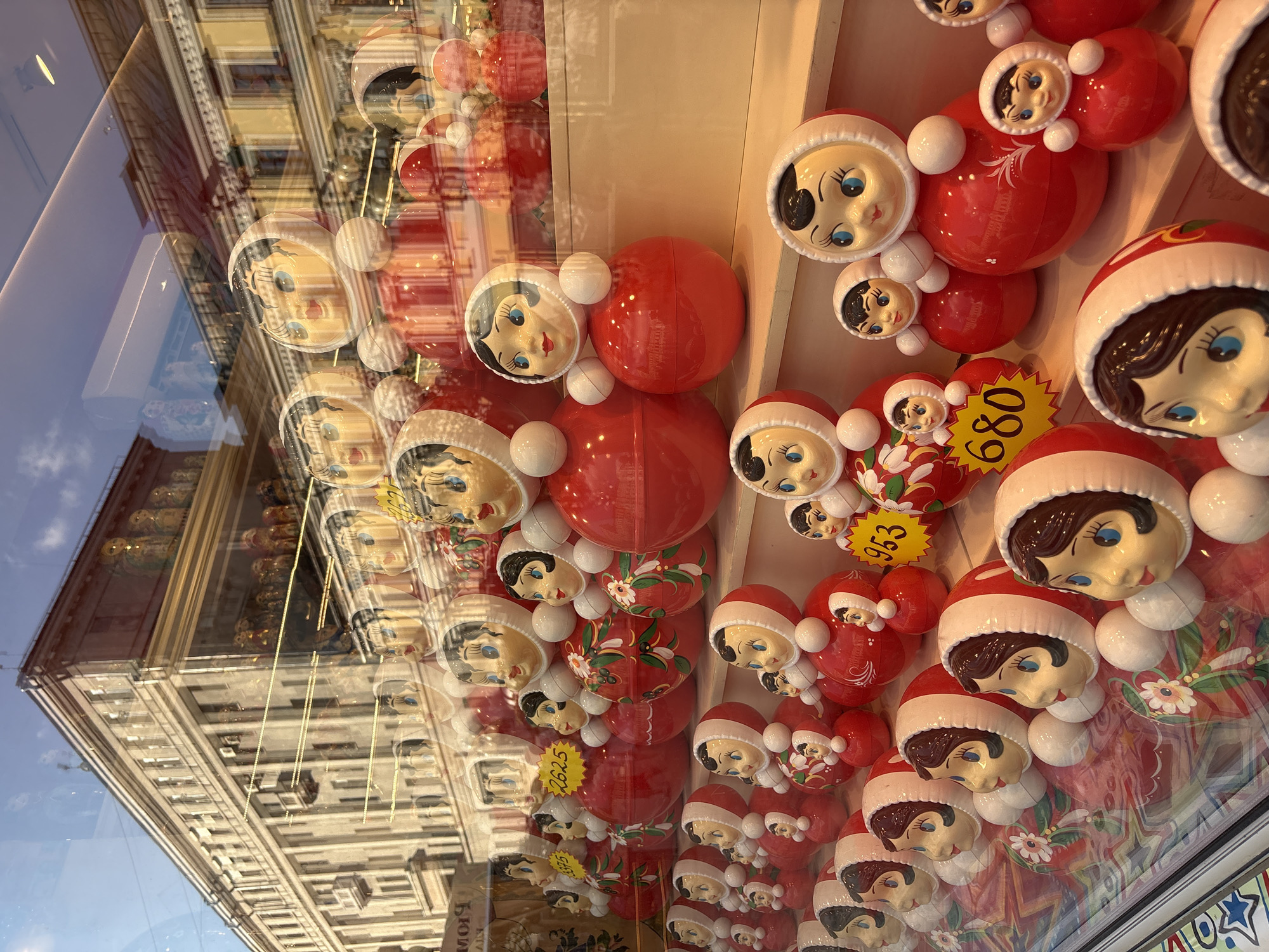The image depicts a display of matryoshka dolls, also known as Russian nesting dolls, in a shop window. The dolls are predominantly red and white, with painted faces featuring blue eyes and rosy cheeks. The dolls are arranged in rows on shelves, with larger dolls in the back and smaller ones in front. Some dolls have price tags attached, displaying numbers such as "953" and "680". The reflection in the window shows a building across the street, appearing to be a multi-story structure with architectural details common in Moscow. The sky above is partially visible with clouds. The overall scene suggests a retail environment showcasing traditional Russian souvenirs.
