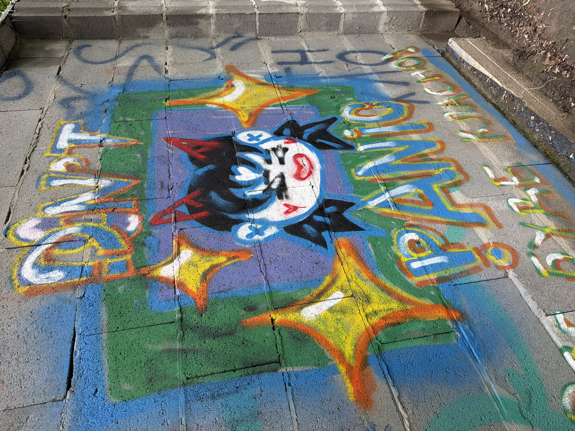 Graffiti adorns a concrete block wall in Kyiv, Ukraine. The wall is painted with bright colors, featuring a cartoon character with red horns and large, expressive eyes. The character's face is framed by black hair and a white heart-shaped nose. Above the character, the words "DON'T" are spray-painted in large, stylized letters, outlined in yellow and orange. Below the character, the word "PANIC!" is also spray-painted in large letters, with a blue background and orange and yellow outlines. To the right of "PANIC!", smaller text in Russian reads "КЛАСНО!", meaning "cool" or "great". The wall is cracked in several places, and some older, faded graffiti is visible in the background. The ground in front of the wall is covered with dirt and debris, with a small concrete ledge.