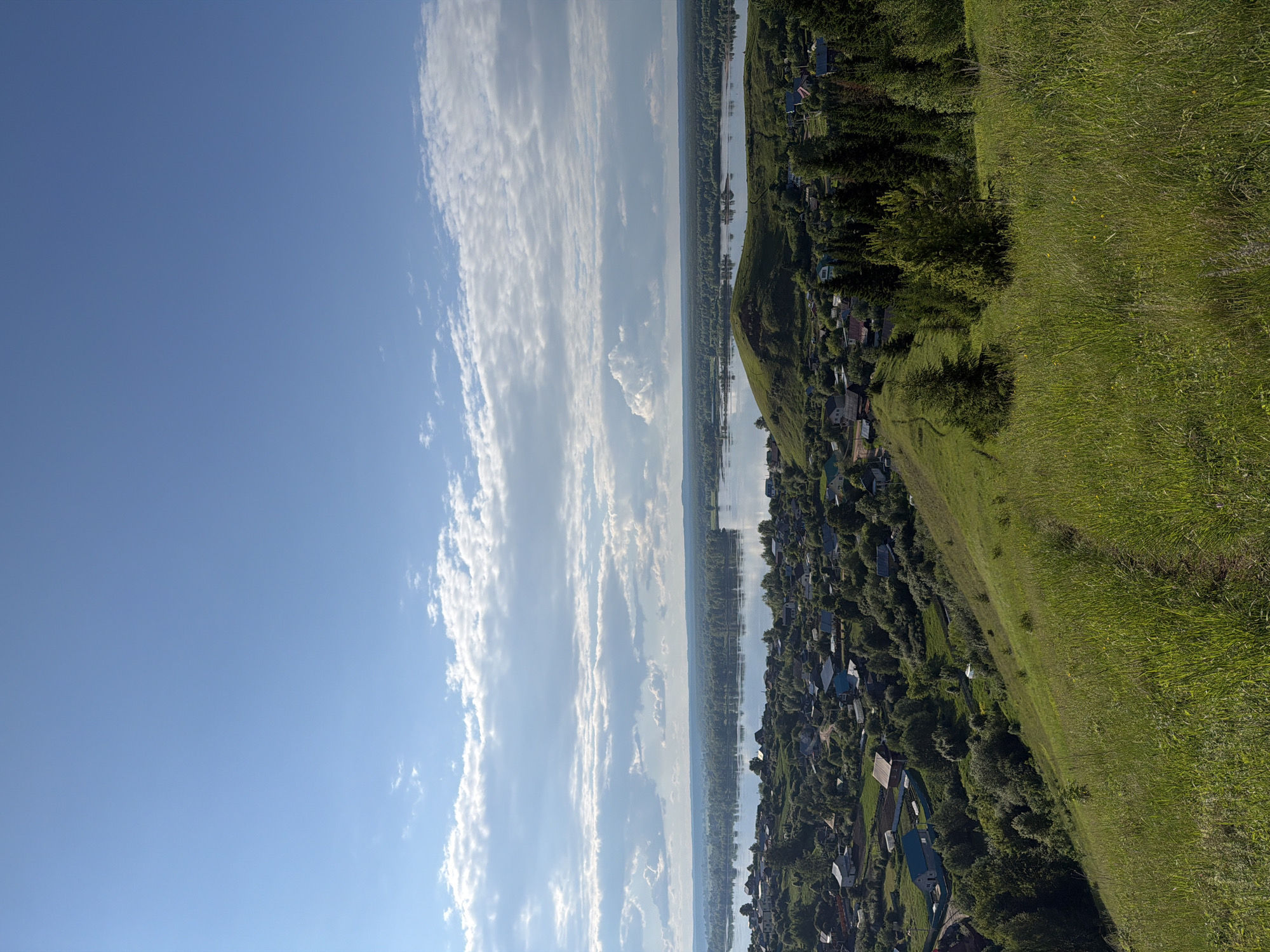 This panoramic image, taken from a high vantage point in Ольховка, Russia, presents a vast landscape under a bright blue sky adorned with long, streaky white clouds. A wide river, reflecting the sky and clouds, flows horizontally across the middle ground of the scene. On the far bank of the river, a distant, unbroken line of dark green forest stretches towards the horizon. On the near bank, a sprawling village composed of numerous houses with varied roof colors (dark, gray, and brown) is visible. These dwellings are interspersed among a dense cluster of lush green trees that extend along the riverbank and up a gentle slope. The foreground, positioned on the right side of the frame, is dominated by a steeply rising hillside blanketed in vibrant, tall green grass. Bright sunlight illuminates the entire scene, indicating a clear, pleasant daytime, likely late morning or early afternoon. The perspective offers a tranquil overview of the residential area, the expansive river, and the natural surroundings. No individuals or specific activities are discernible within the frame. There is no visible text.