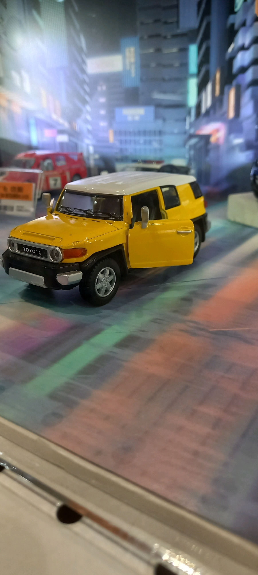 A die-cast model of a yellow and white Toyota FJ Cruiser is displayed on a surface that reflects colorful, abstract patterns. The car is facing left and is partially open with the driver's side door ajar. The background is a blurred, urban cityscape with tall buildings and neon lights, suggesting a nighttime setting. The overall impression is of a display or exhibit, possibly in a store or a showroom. There is no indication of Samarinda, Indonesia in the image.