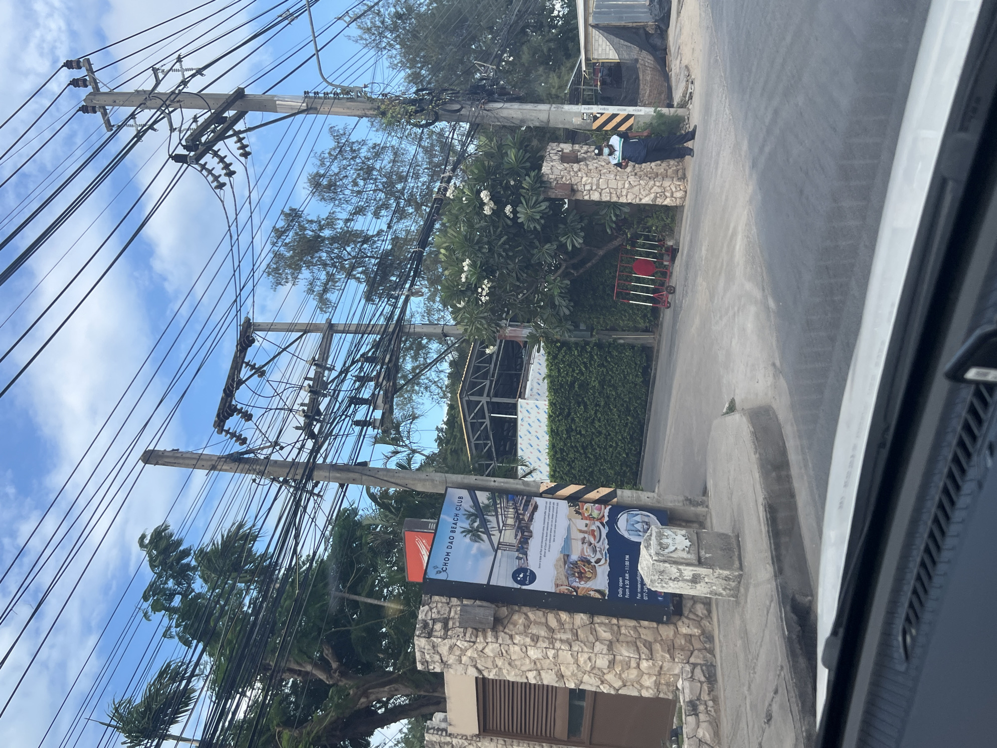In a city location of Unknown, Unknown, a security guard stands near a stone wall entrance to "Chom Dao Beach Club." The guard is wearing a dark blue uniform with a lighter blue stripe and a mask. The entrance is adorned with a sign advertising the beach club, which displays images of food and its operating hours. A red portable barrier is situated near the entrance. Above the scene, numerous tangled electrical wires are suspended from wooden poles against a backdrop of a blue sky with scattered clouds. Trees, including palm trees, and some tropical foliage surround the area. The foreground shows a portion of a car's dashboard and windshield.