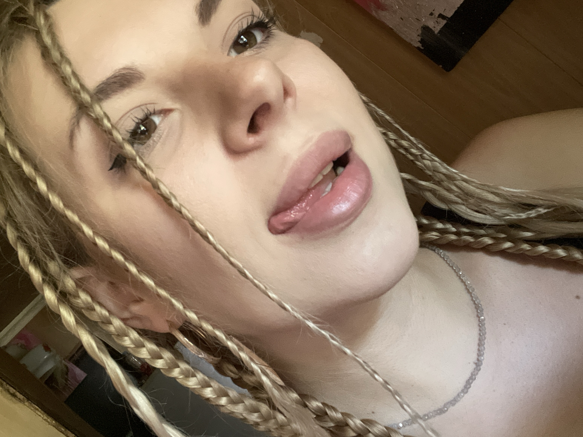 A close-up, low-angle selfie shows a young woman with blonde braided hair in Shcherbinka, Russia. She is wearing a beaded necklace. Her tongue is slightly extended, and she has on lip gloss and eyeliner. The background is out of focus, but appears to be an indoor setting with wooden flooring.