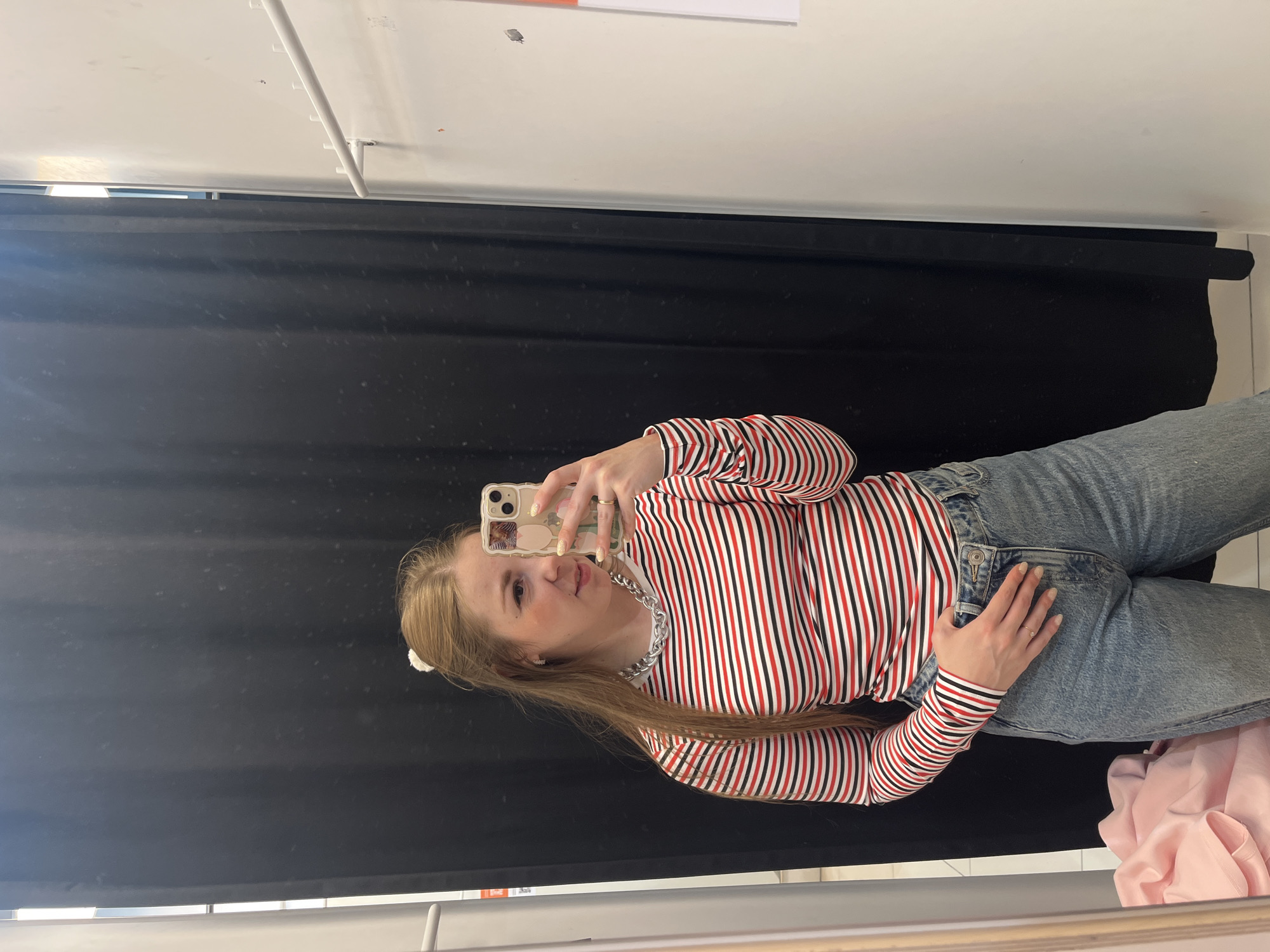 A young adult female is depicted in a mirror selfie taken within what appears to be a retail store fitting room. She wears a long-sleeved top with alternating red, white, and black horizontal stripes, paired with light blue, loose-fitting denim jeans. Her light-colored hair is styled partially up with a white scrunchie, while the remainder flows over her left shoulder. She sports a chunky silver chain necklace, a small ring on her left ring finger, and small earrings. Her right hand holds a light-colored smartphone with a patterned case, angled to capture her reflection. Her left hand rests on the waistband of her jeans. Behind her, a dark curtain acts as a privacy screen. To the right of the reflected image, a white wall is visible, equipped with a white horizontal hanging rack featuring multiple pegs. A pile of light pink fabric is present on the floor in the bottom left corner of the reflected image. The lighting is bright and even. The location is an indoor commercial space in the city of Unknown, Unknown.