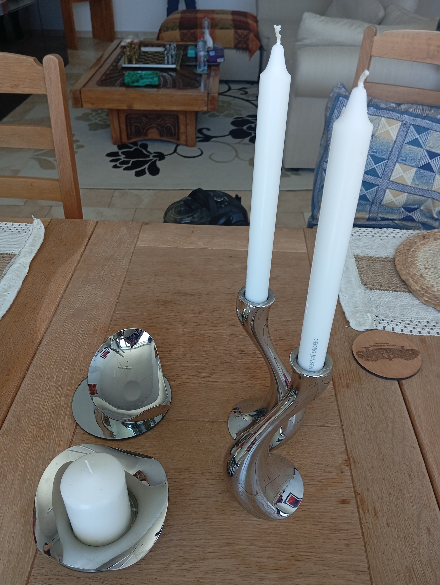 Two tall white taper candles are seated in a modern, chrome-finished, sculptural candelabra, which rests on a light-colored wooden table. To the left, a smaller, similarly styled chrome holder contains a single white pillar candle. The table is set in what appears to be a dining or living area with a patterned rug visible in the background, a wooden coffee table with items on it, and furniture including chairs and a sofa. The overall scene suggests a home environment.