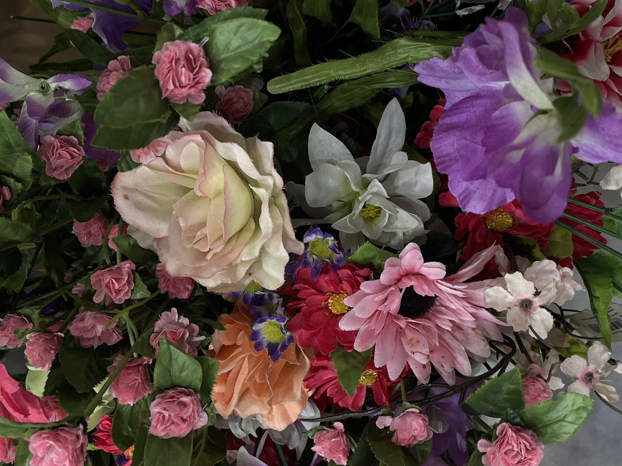 This is a close-up, eye-level, vertical, still shot of a vibrant bouquet of artificial flowers. The flowers are densely packed, showcasing a variety of colors including shades of pink, peach, red, white, purple, and green foliage. Prominent flowers include a large pale pink and yellow rose, a bright orange carnation-like bloom, a large pink daisy with a dark center, a cluster of small pink roses, and a large purple flower in the foreground. The lighting is even, with no shadows indicating the light source. The background is a dark, indistinct blur of more flowers and greenery. There are no people or animals visible in the image. There is no discernible information or context within the image that would suggest a specific location in Davao City, Philippines.
