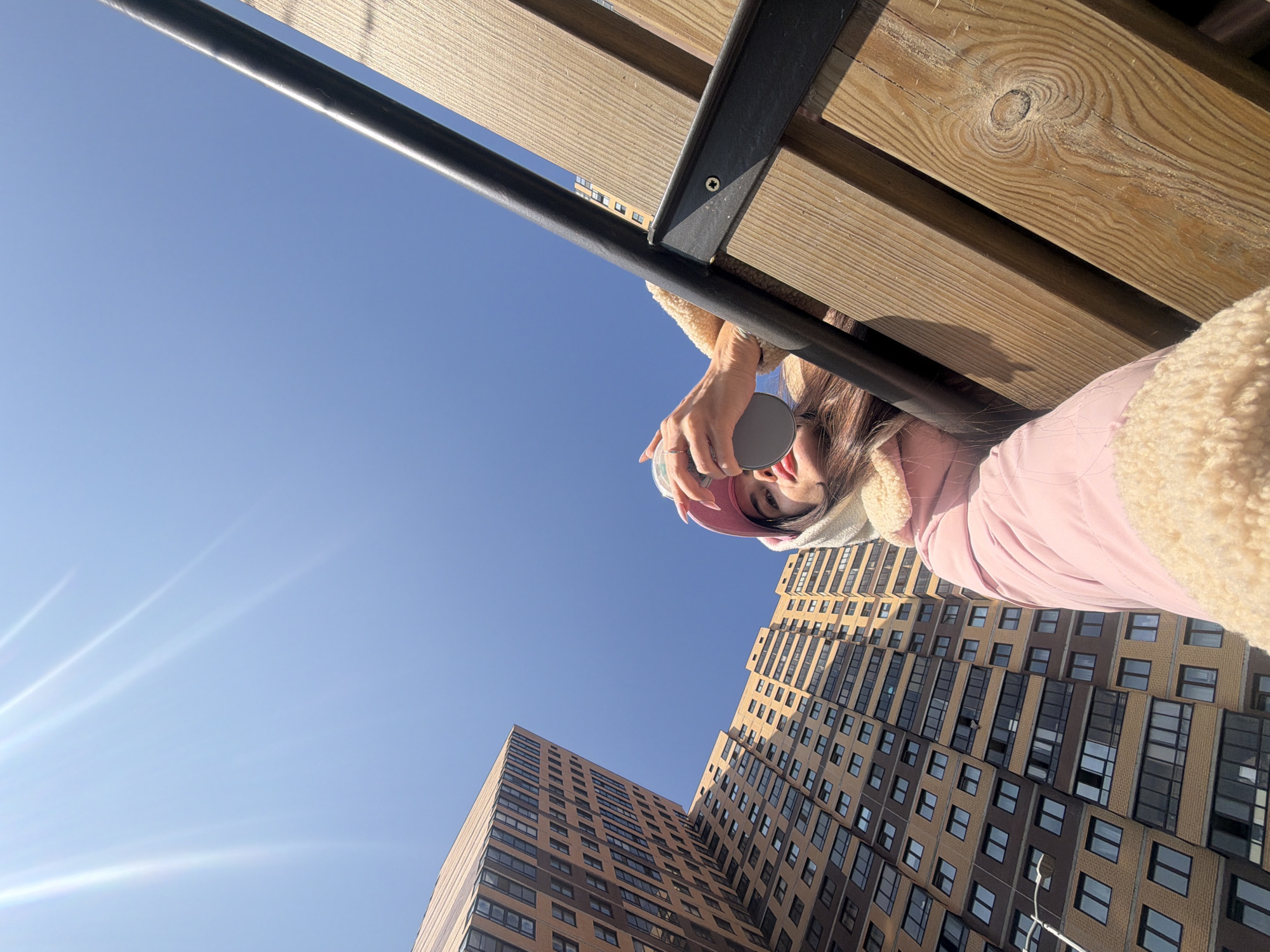 A low-angle shot looking upward at a woman sitting on a wooden bench in front of two tall buildings. She is holding a grey cup with a lid. She wears a pink jacket, a white fluffy collar, and a pink hat. The buildings are light brown with dark windows. A blue sky is visible behind the buildings and above the bench. The bench has wooden slats with a black metal frame. The image was taken in Saint Petersburg, Russia.