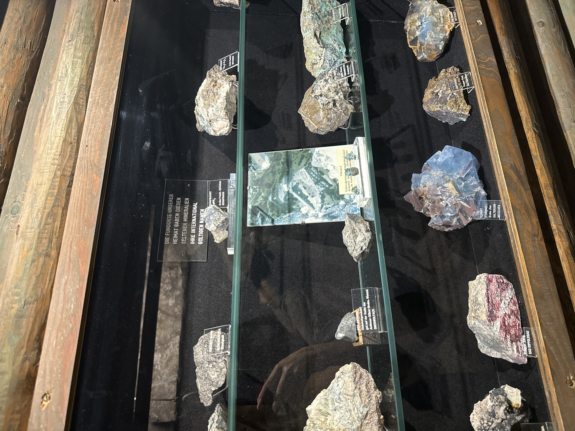 The image displays a museum exhibit featuring various mineral specimens in a glass case. The exhibit is labeled "DIE FUNDORTE UNSERER HEIMAT GABEN DIESEN SELTENEN MINERALIEN IHRE INTERNATIONAL GELTIGEN NAMEN". The display is organized on multiple shelves, with each mineral specimen resting on a small clear stand with a label. Some of the labels include names such as "GED. KUPFER" and "FLUORIT". The background of the display case is black, and the lighting highlights the textures and colors of the minerals. Wooden beams frame the top of the display. The location is specified as Schonach im Schwarzwald, Germany, indicating this is likely a local museum or exhibition.