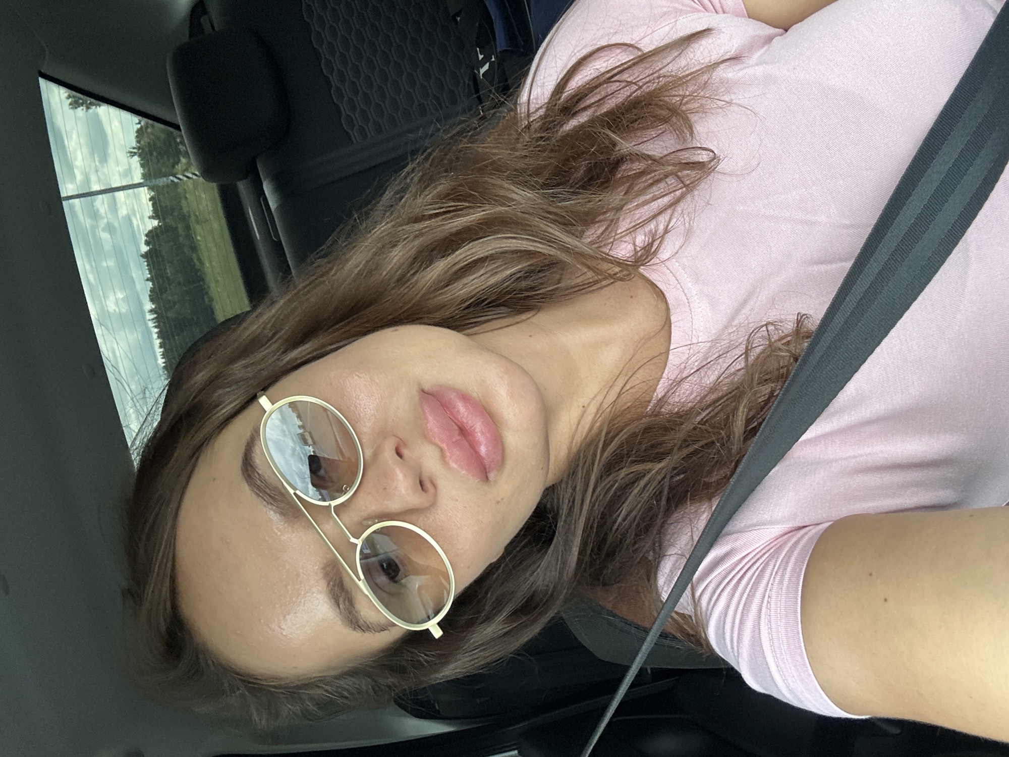 A woman is taking a selfie in a car. She is wearing a pink t-shirt and sunglasses. The interior of the car is visible, with the back seat and headrests in the background. The window shows a rural landscape with trees and a cloudy sky. The image is cropped to focus on the woman's face and upper body. The location is not Karsy, Russia; it is a generic rural landscape.