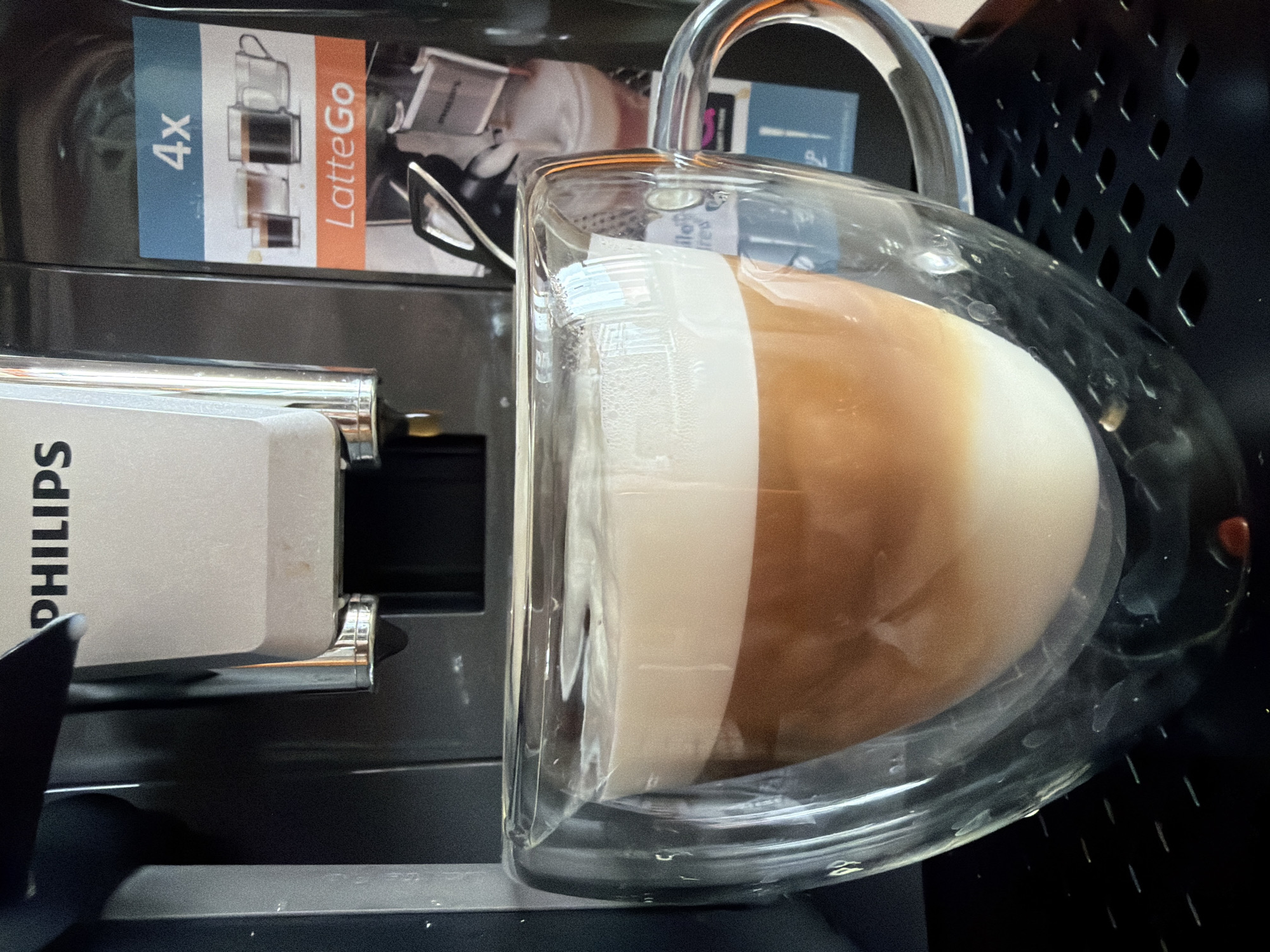 This image, captured from a rotated perspective, presents a close-up view of a freshly prepared, layered coffee drink in a clear, double-walled glass mug. The drink distinctly shows a darker coffee layer at the bottom and a thick, frothy white milk layer on top, characteristic of a latte or cappuccino. Small bubbles are visible within the milk foam, and the mug has a clear handle.
Adjacent to the mug, a portion of a coffee machine is visible. Its silver-colored body displays the brand name "PHILIPS" printed vertically. On a darker section of the machine, a label is affixed, showing "4x" on a blue background and "LatteGo" on an orange background. This label also includes small graphical representations of different cup sizes. The area behind the mug and coffee machine is dark, with a textured, possibly perforated surface on the right. The overall scene suggests an indoor setting, likely a kitchen or a coffee preparation area, situated in Chaikovsky, Russia.