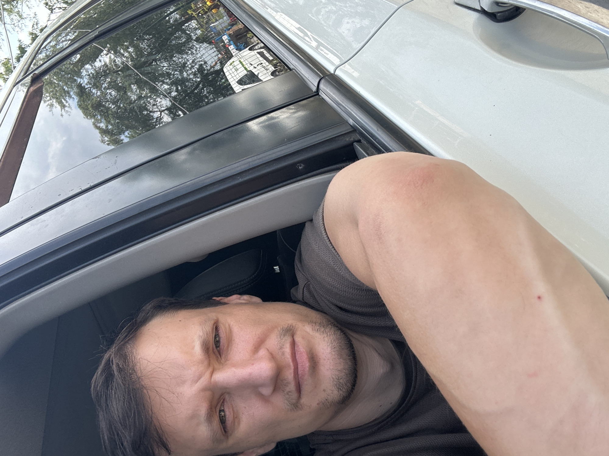 A man with short brown hair and stubble is leaning out of a car window, with his left arm resting on the exterior of the vehicle. He is wearing a dark t-shirt. The car appears to be white. Through the car window, trees and a cloudy sky are visible, as well as a white van with a green logo. The scene is outdoors, with hints of a city environment in the background, possibly Shatava, Ukraine.