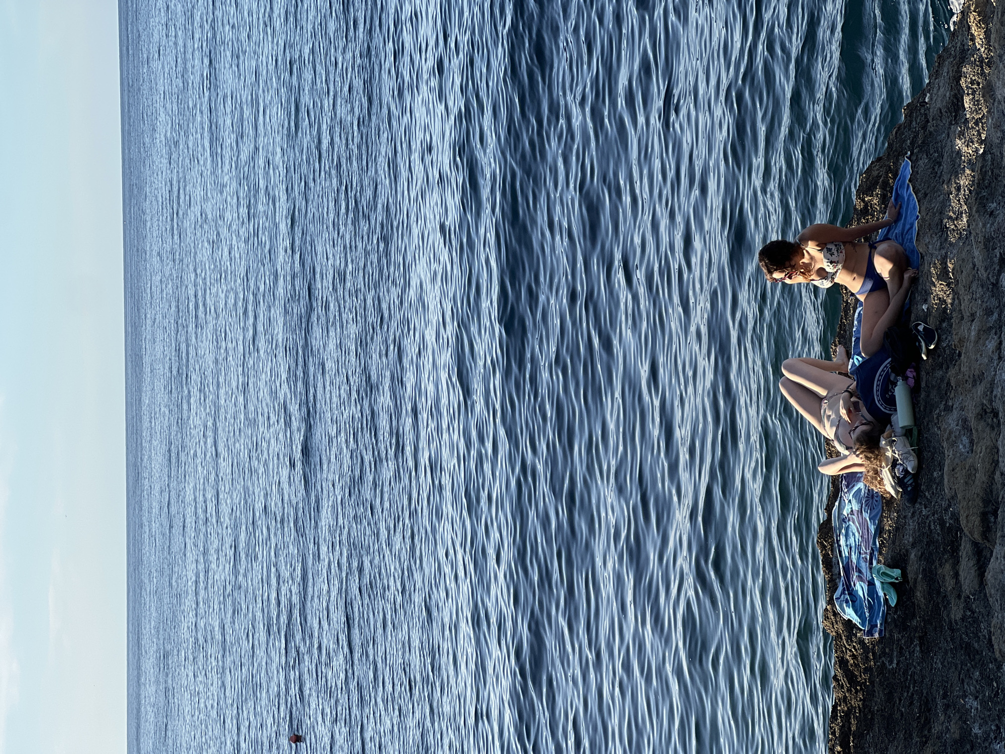 The image depicts a coastal scene in Sperone, Italy, featuring two individuals relaxing on a rocky shoreline adjacent to the sea under a clear sky. The horizon line is distinct, separating the light blue sky from the dark blue, rippled ocean. In the water, a small red buoy is visible on the left side, and a swimmer's head is partially submerged further out in the upper-left quadrant.
On the dark, rugged rocks in the foreground, two women are present. The woman on the left is lying on her back, facing upwards, on a multi-colored (blue, purple, white) towel. She is wearing a light-colored bikini and has curly reddish-brown hair. Beside her, a pair of light blue/green sandals, light-colored sneakers, and a dark piece of fabric or bag are placed. The woman on the right is seated upright on a blue towel, looking towards the right side of the frame, away from the camera. She is wearing a white floral-patterned bikini top and dark blue bikini bottoms, with sunglasses resting on her head. Near her feet are a dark bag and a pair of black sneakers. Both individuals appear to be sunbathing or relaxing by the sea.