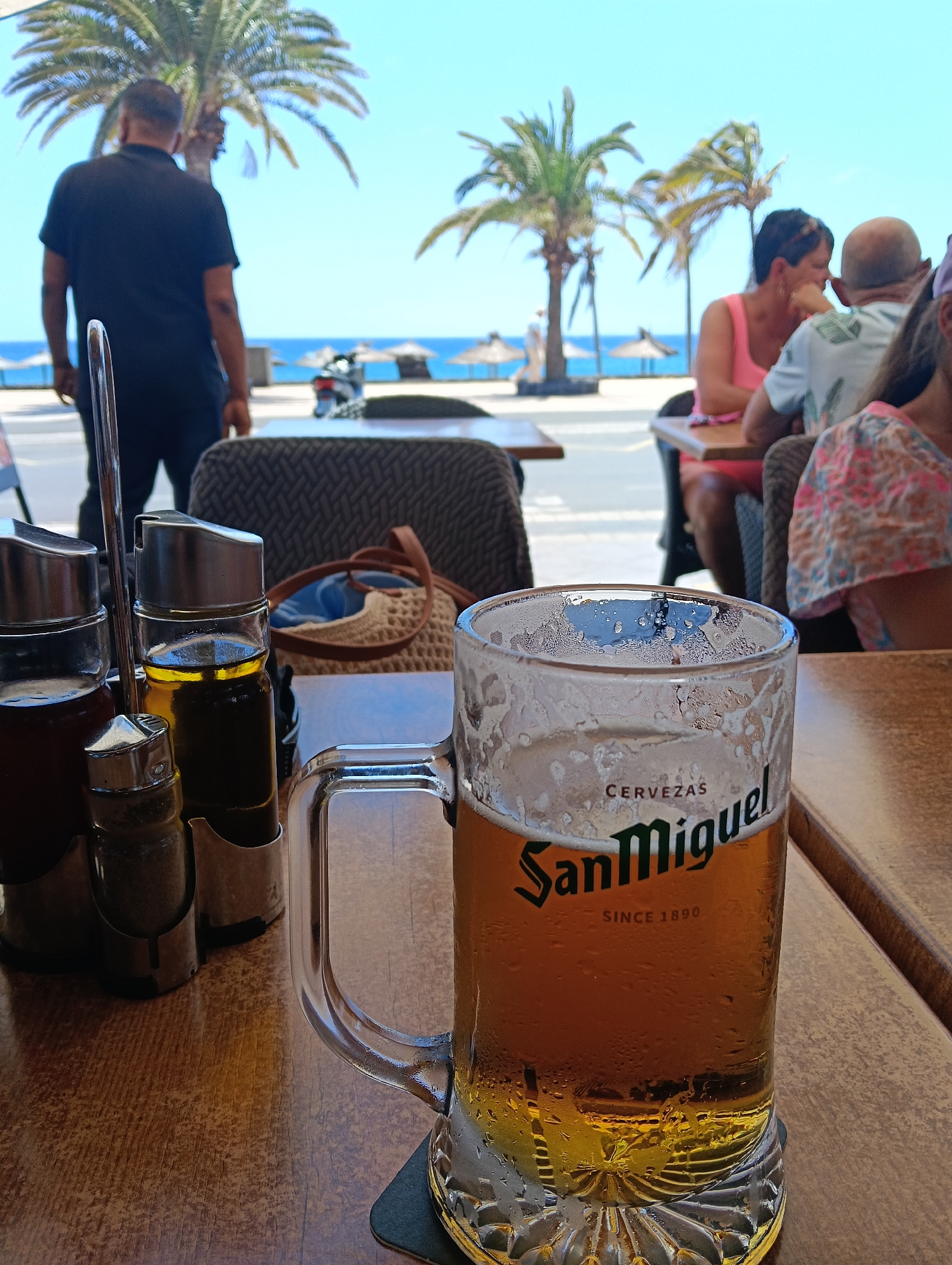 The image presents a sunny, daytime scene from an outdoor cafe or restaurant in Puerto Del Carmen, Spain, with the ocean visible in the background.
In the foreground, a wooden table holds a clear glass mug of San Miguel beer, which has condensation on its surface, indicating it's cold. The mug clearly displays "CERVEZAS San Miguel SINCE 1890". A black coaster is beneath it. To the left, a metal caddy holds four condiment bottles, including one with red sauce, one with olive oil, and shakers likely for salt and pepper.
Beyond the table, a diverse group of people are seen. To the left, a man in a black polo shirt stands with his back to the viewer, looking towards the sea. To the right, several individuals are seated at other tables, including a woman in a pink top engaged in conversation with a bald man in a light-colored patterned shirt. Part of a child or another person in a floral-patterned top is also visible. Their activity suggests casual dining or socializing.
The background reveals a vibrant coastal setting. A street or promenade separates the cafe from the beach. Several tall, green palm trees are scattered throughout the scene. The sky is a bright, clear blue, meeting a tranquil azure ocean. On the beach, a few straw-thatched umbrellas are visible. A scooter is parked near the road. The overall atmosphere is relaxed and indicative of a holiday destination.