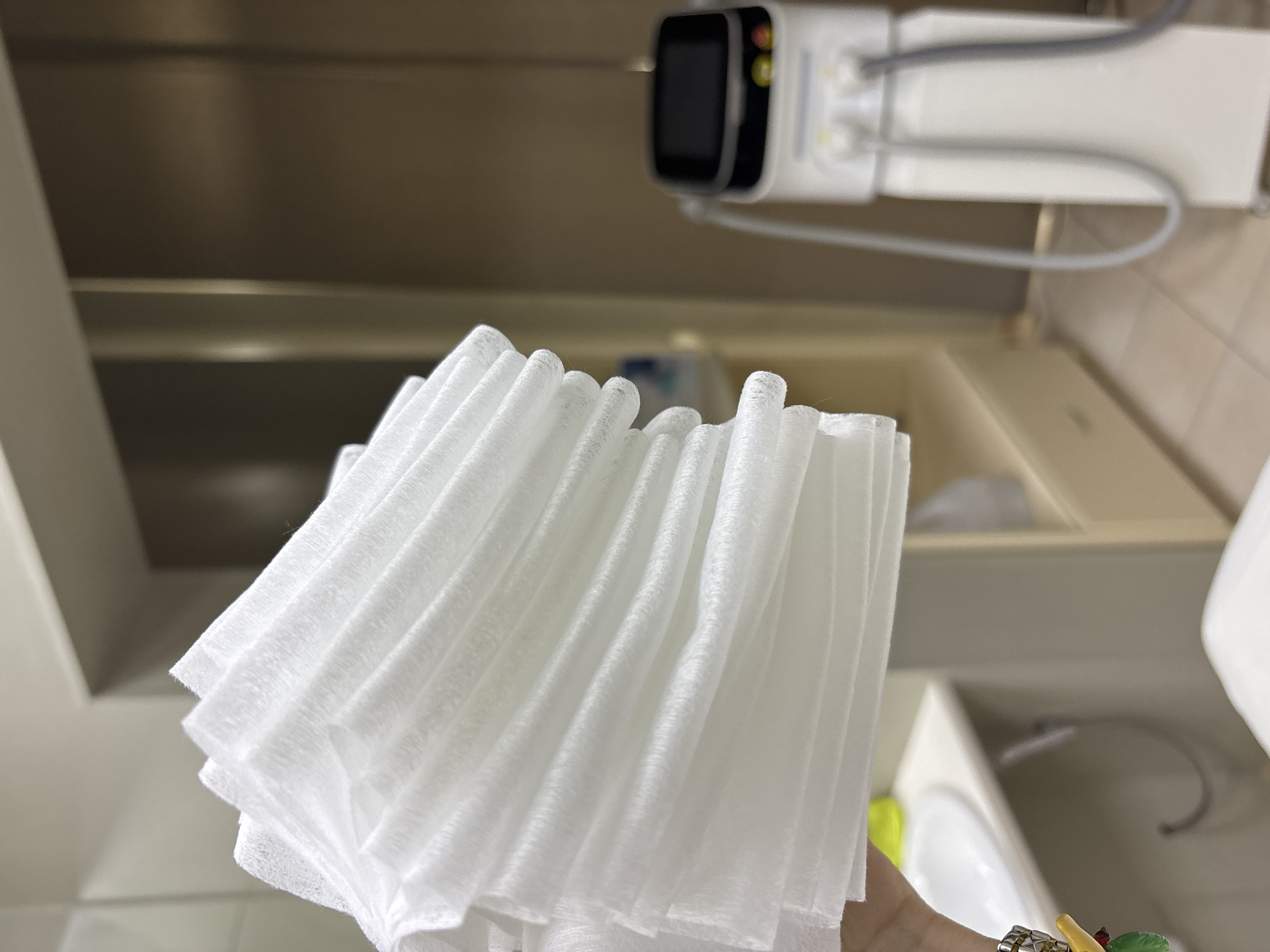 A close-up shot displays a stack of white, possibly disposable, facial cloths. The cloths are neatly arranged with slightly uneven edges. In the background, a white electronic device with a screen is mounted above a cabinet. The surrounding environment suggests a bathroom or medical setting, with glimpses of a sink and countertop visible. The specific location appears to be indoors.