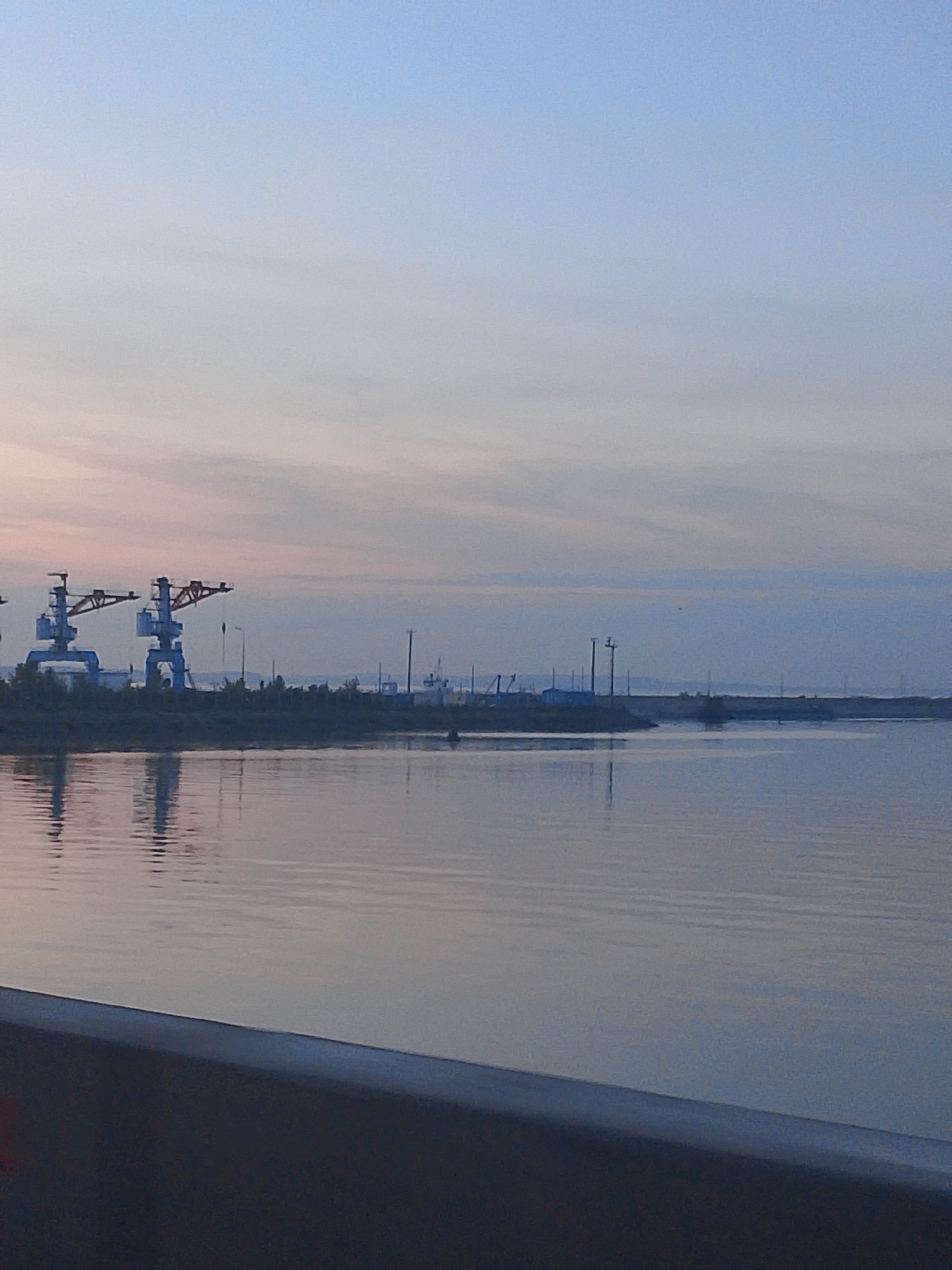 The image depicts a waterfront scene in Chaikovsky, Russia. In the background, several port cranes are visible along the shoreline. Power lines and distant buildings are also discernable in the background. The foreground is dominated by a body of water, which reflects the sky and shoreline elements. The sky is light blue with hints of orange and pink at the horizon, indicating either sunrise or sunset. A portion of a railing or bridge structure is visible at the bottom of the image.
