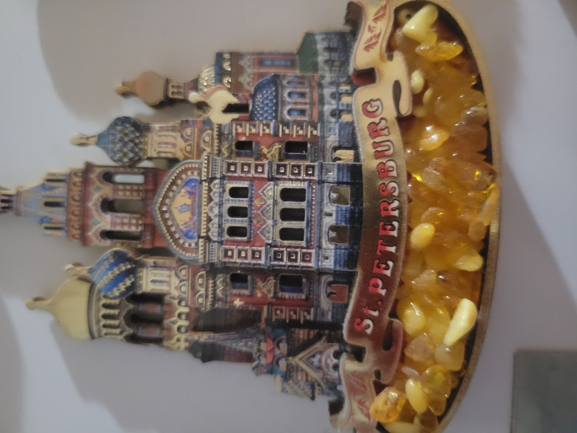 The image displays a souvenir magnet depicting the Church of the Savior on Spilled Blood in St. Petersburg, Russia. The magnet is shaped like the church and features intricate details of its architecture, including its colorful domes and ornate facade. The words "ST. PETERSBURG" are clearly visible on a banner across the front of the magnet. The lower portion of the magnet is filled with small, yellow, translucent pieces that resemble amber. The magnet is affixed to a light-colored, textured wall, with a shadow cast on the upper left side of the wall. There are no people or actions depicted in the image. The location of Belaazyorsk, Belarus is not relevant to this image.