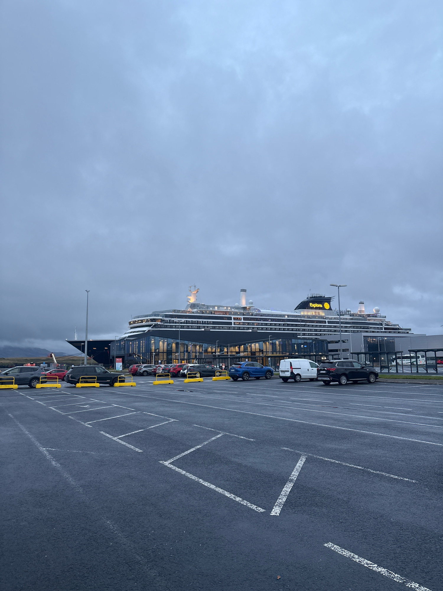 The image depicts a cruise ship docked at a terminal in Reykjavik, Iceland. The sky is overcast and gray, suggesting a cool or rainy day. In the foreground, an asphalt parking lot with painted white lines for parking spaces is visible. Several cars, including SUVs and a white van, are parked in the lot. Yellow barriers are placed in front of some of the parking spaces.
The cruise ship, identified by the illuminated "Explora" logo on its funnel, is a large vessel with multiple decks and white superstructure. It is docked alongside a modern building with large glass windows, which appears to be the cruise terminal. The building has a sleek, contemporary design. Mountains or hills are visible in the background on the left side of the image, shrouded in a misty haze. The overall scene suggests a port facility in a northern climate, likely during a cloudy day. There are no people visible in the image.