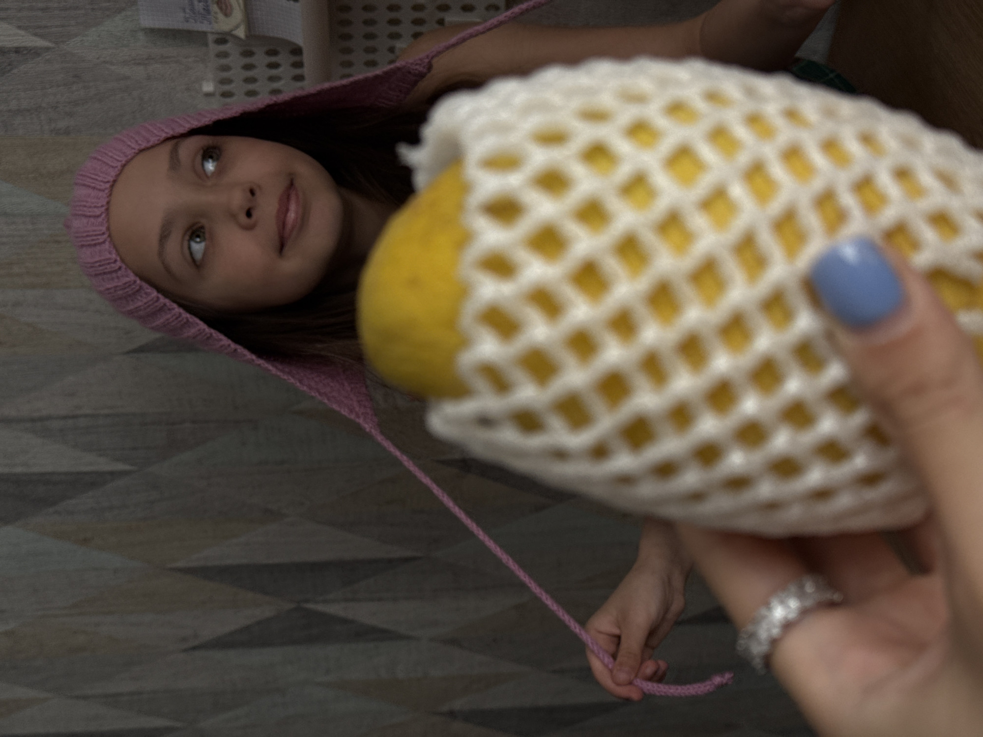 A young girl wearing a pink bonnet and looking up is partially visible. A yellow fruit is in a white protective mesh. A hand is holding the fruit. The hand has a ring and a blue painted fingernail. The background shows a grid pattern, and the floor has a patterned design. The location is not discernible, and Votkinsk, Russia, cannot be confirmed.