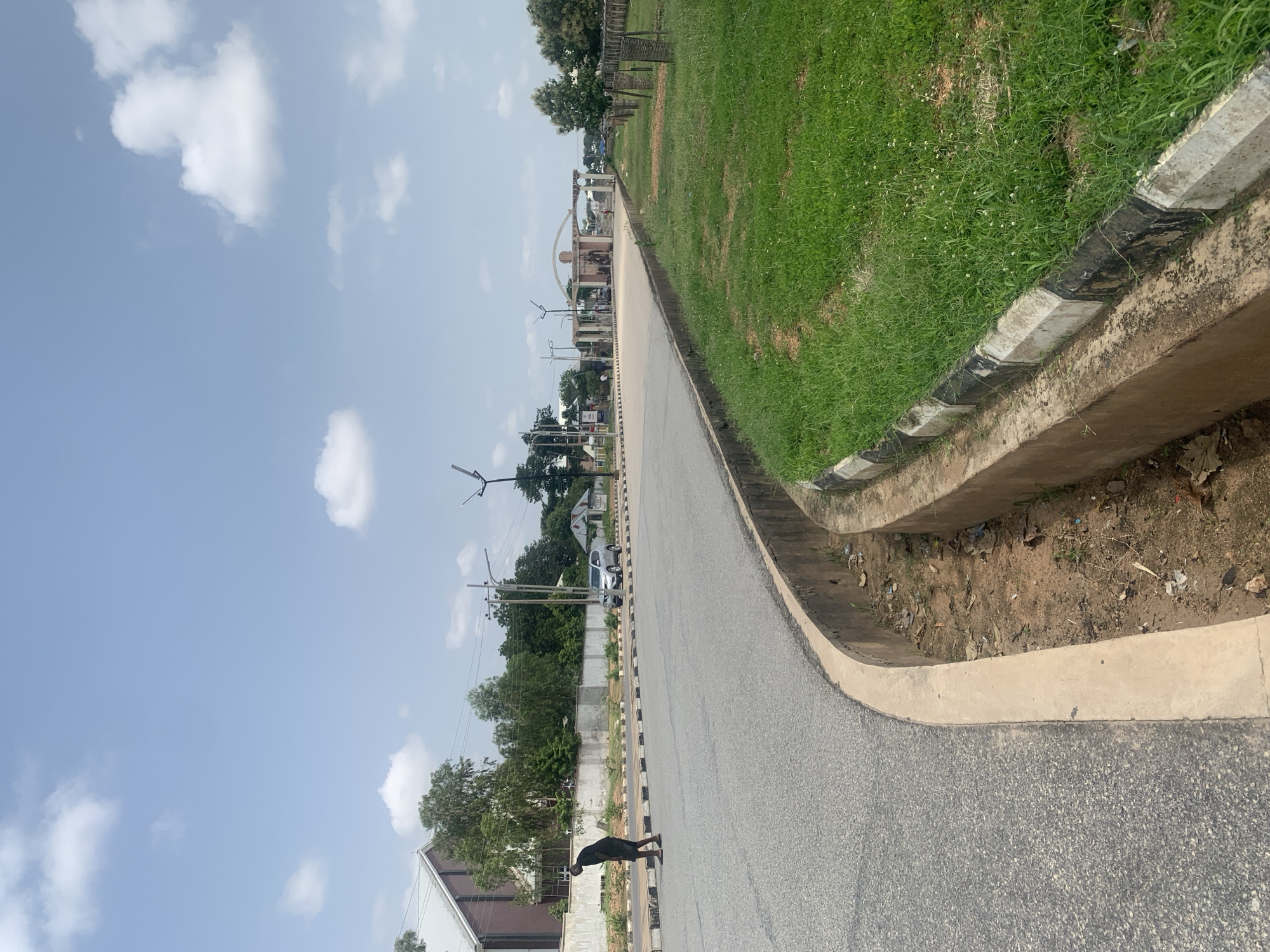 A medium shot captures a street scene on a clear day in Jalingo, Nigeria. A lone person, appearing to be a male dressed in dark clothing, walks across the road from left to right. The road is paved and curves to the right. A concrete drainage ditch runs along the right side of the road, filled with dirt and some debris. Adjacent to the ditch is a patch of well-maintained green grass. In the background, a silver sedan is parked on the side of the road, and further down, a large, arched gateway structure is visible, flanked by columns and appearing to be an entrance to a facility. Trees and buildings are also present in the distance. The sky is blue with scattered white clouds.