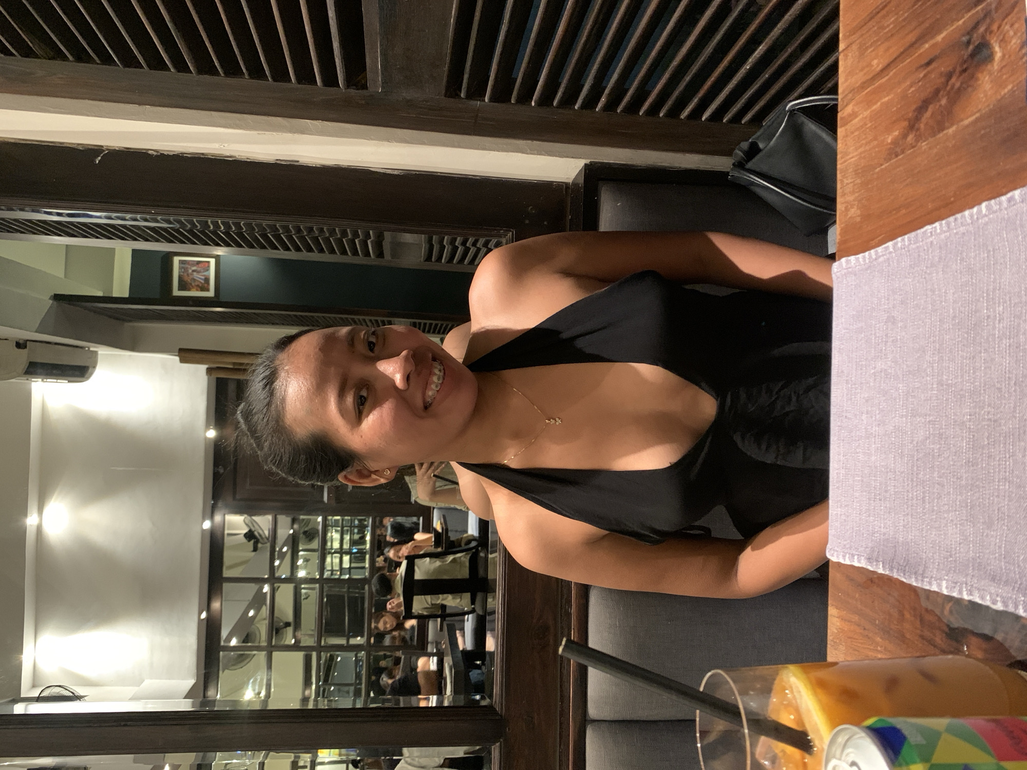 A woman with dark hair and a black top is seated at a table in a restaurant in Kuta, Indonesia. She is smiling and facing the camera, and a straw is in her drink. Other people are visible in the background behind her in the reflection of the large windows, indicating a busy restaurant setting. The restaurant has a wooden-slatted ceiling, and the table is also made of wood, with a gray placemat on it.