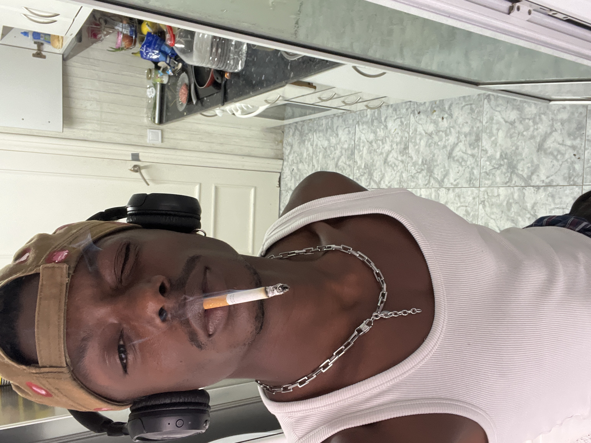 An individual is captured in a close-up portrait, oriented in an indoor setting that appears to be a kitchen or utility area. The subject is a person with dark skin, wearing a white ribbed tank top and a silver chain necklace. A light brown baseball cap with what looks like small reddish or pink details is worn on their head, and black over-ear headphones rest around their neck. They have a lit cigarette in their mouth, with a plume of smoke visible around their face. Their eyes are partially closed, suggesting a relaxed or contemplative expression, possibly influenced by the smoke.
The background reveals a cluttered domestic environment. Behind the individual, a countertop is visible, laden with various items including a large plastic water bottle, cooking pots or pans, and several plastic bags with contents. The wall behind the countertop features horizontal white paneling, and further to the right, a greyish-white tiled wall with a marbled pattern can be seen. Below the counter, there are white cabinets with handles. A white door with paneling and a metallic handle is also partially visible on the left side of the frame. The lighting suggests an indoor setting, with no specific indication of the time of day. No discernible text is visible on any objects or surfaces.