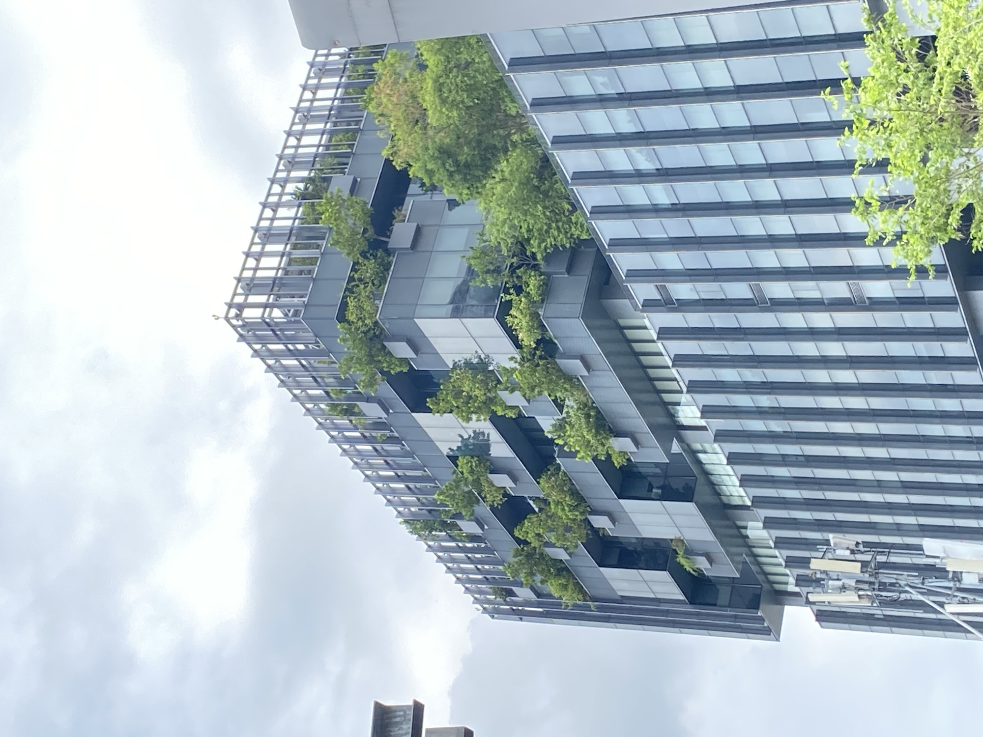 A modern skyscraper with a distinctive architectural design featuring integrated greenery stands against a cloudy sky. The building's facade is composed of reflective glass panels and staggered geometric shapes, with several tiers hosting various trees and plants. The structure's steel framework is visible, adding to the modern aesthetic. The image's perspective is from below, looking upwards at an oblique angle, emphasizing the building's height. The building is located in the city of Bangkok, Thailand.