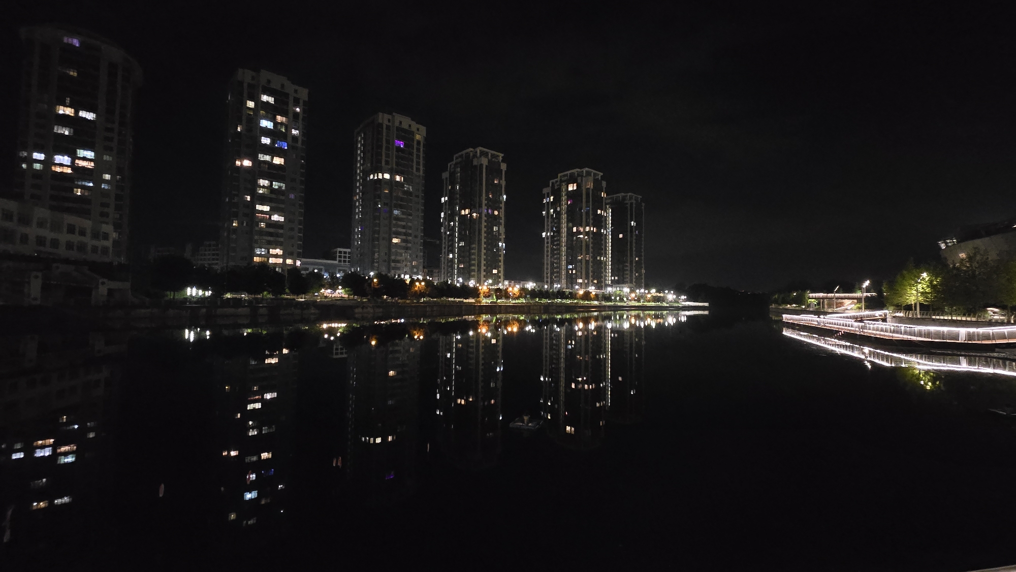 The image displays a night scene along the Rusaniwka Canal in Kyiv, Ukraine. Six tall, modern residential buildings with illuminated windows dominate the left and central portions of the frame, their structures and lights clearly reflected in the still, dark water below. The building on the far left features a rounded silhouette, while the subsequent five buildings are rectilinear. Window lights within the buildings present varying colors, including white, yellow, and faint purple. Along the right bank of the canal, a curving promenade or walkway is brightly illuminated by linear white lights, which also cast distinct reflections on the water. A sparse line of trees is visible along the banks between the buildings and the water, with some individual trees on the far right also illuminated. The sky above is dark and featureless. No individuals, vehicles, or explicit actions are present within the captured scene.