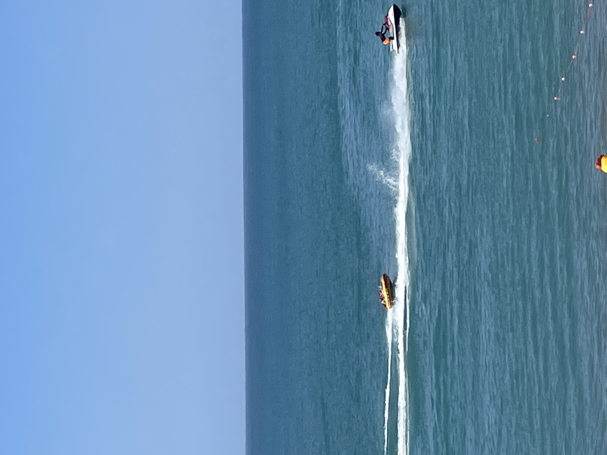 The image depicts a bright, sunny day at a beach in Giachrypsh, Abkhazia. The sea is a vibrant blue, with gentle waves. In the foreground, the edge of the beach is visible with some small buoys connected by a string. Two watercraft are active on the sea. On the right, a person is riding a jet ski, creating a white spray of water. To the left of the jet ski, an inflatable tube, also known as a "banana boat," is being towed, with several people riding on it. The sky is a clear, pale blue.
