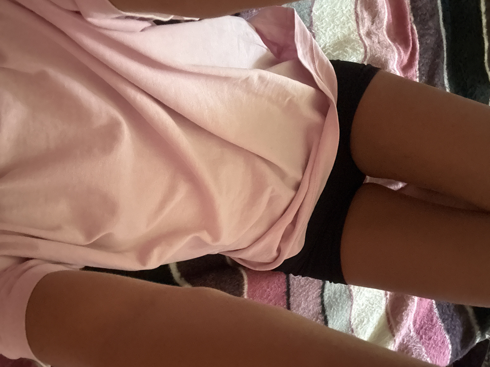 The image shows a close-up view of a person's torso and legs, lying down. They are wearing a pale pink t-shirt that drapes over their black shorts. The person appears to be lying on a striped blanket with colors including white, pink, and dark purple. The lighting suggests an indoor setting, and the angle of the shot is from above, looking down at the person. There is no visible text in the image. The overall impression is one of casual relaxation or rest.