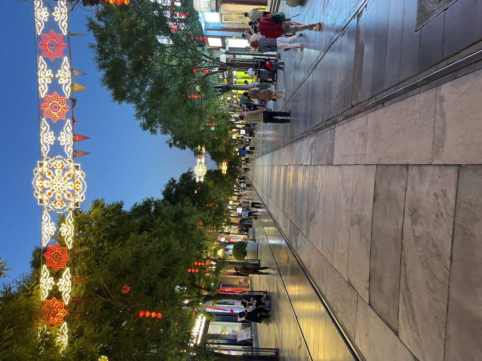 The image shows a pedestrian street in Dongcheng District, China, at dusk. The street is lined with trees adorned with red lanterns and decorative light fixtures, resembling stylized floral patterns. People are seen walking and standing along the street. The street surface is made of stone slabs with metal strips running along the sides. The sky is a gradient of blue.