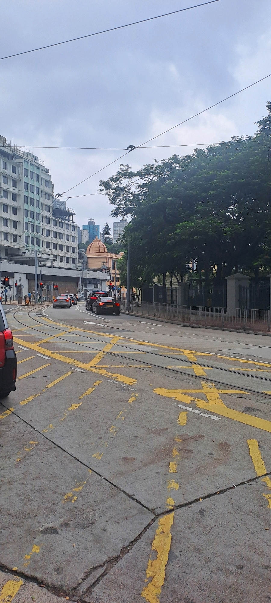 This is a street-level, vertical, medium shot of a cloudy day in Hong Kong, China, likely a tramway intersection in an urban area. The foreground shows the road surface with prominent yellow markings, including crossed lines and diagonal stripes, indicating a traffic junction or hazard zone. Tram tracks are clearly visible embedded in the asphalt.
In the middle ground, several cars are present, including a dark-colored sedan with its rear license plate visible, a silver sedan, and a dark SUV. Traffic lights are visible on poles, with some indicating red. Pedestrians, appearing as small figures, are seen on the sidewalk in the distance.
The background features a large, multi-story white building with numerous windows. To the right of this building, a large tree with dense green foliage dominates the scene. Further in the background, more buildings of varying heights and architectural styles are visible, including one with a distinctive domed roof. Overhead, electrical wires are strung across the frame, typical of an urban environment with a tram system. The overall impression is a busy city street under an overcast sky.
