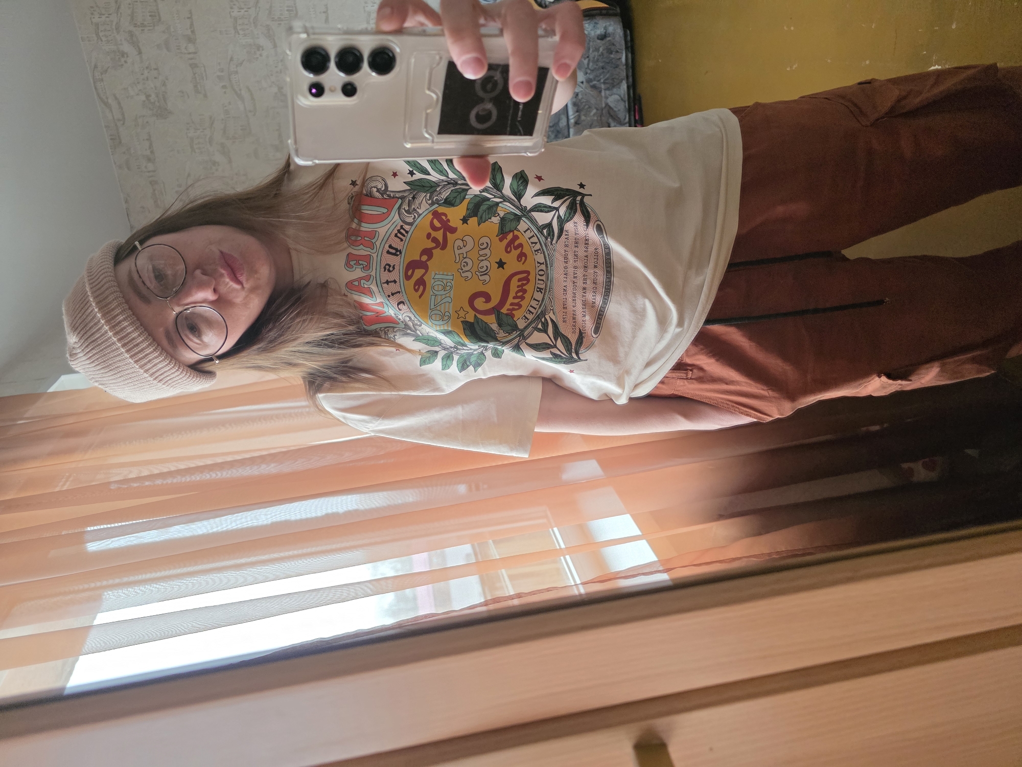 The image appears to be a selfie taken by a person with light brown hair, wearing glasses and a pink beanie. They are wearing a white t-shirt with a graphic print and brown trousers. The person is holding a smartphone with a transparent case. The background consists of a window with orange blinds and part of a wooden cabinet. The image is taken from a slightly tilted angle and the text on the t-shirt is mirrored due to the selfie perspective. There is not enough information to determine the location in Chaikovsky, Russia.