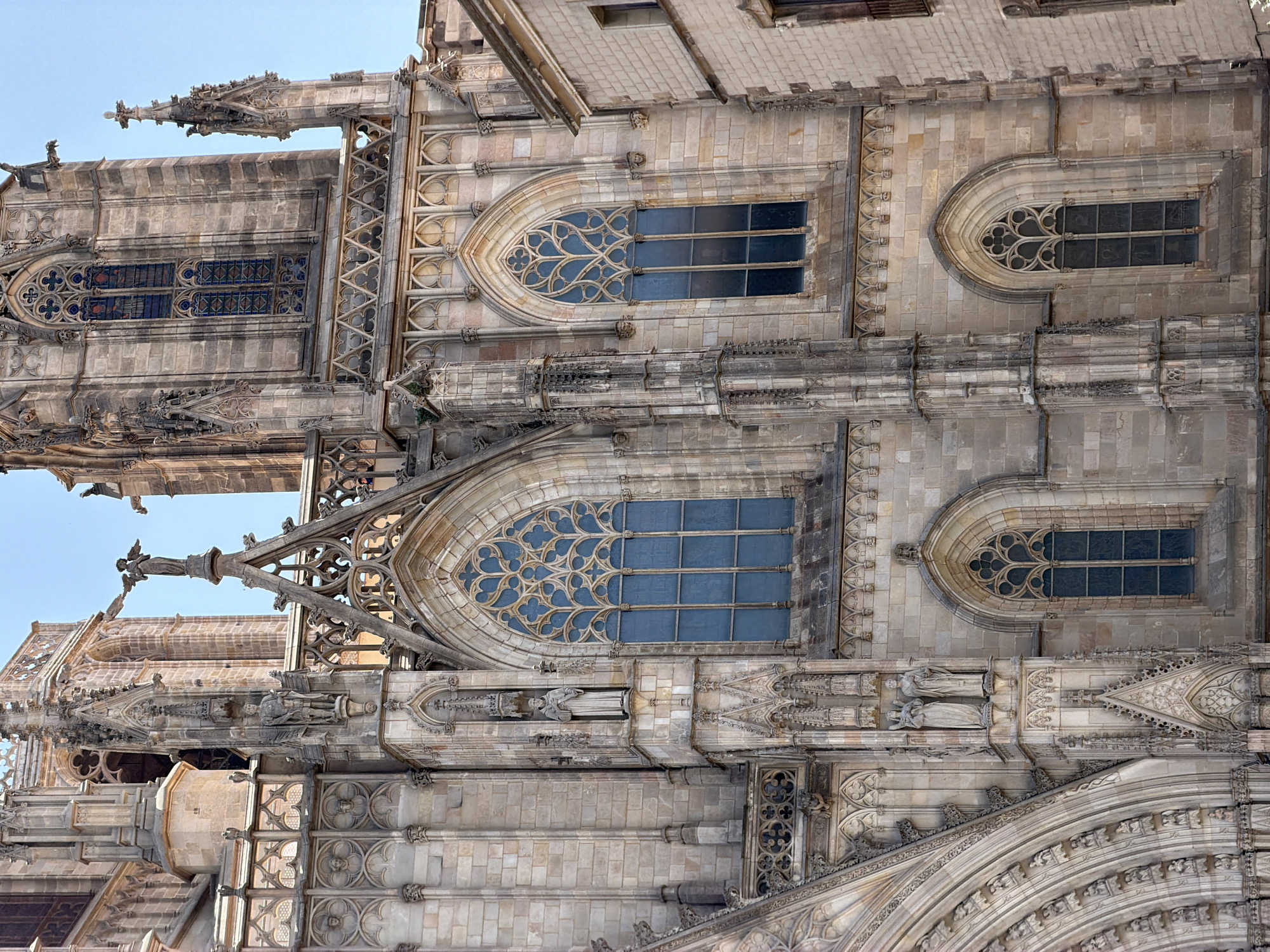 This is a photograph of the exterior of the Barcelona Cathedral, also known as the Cathedral of the Holy Cross and Saint Eulalia, located in the Gothic Quarter of Barcelona, Spain. The image focuses on the intricate architectural details of the cathedral's facade, particularly its Gothic style.
The facade is constructed of light-colored stone, with prominent features including tall, arched windows with elaborate tracery and stained glass, pointed spires, and detailed carvings. Several statues are visible, placed within niches or on architectural projections. One statue appears to be of a robed figure, possibly a saint or a religious icon, standing on a pedestal. Another statue depicts a winged figure, an angel, at the apex of a pinnacle. The windows are tall and narrow, with multi-paneled glass that appears to be a deep blue or purple hue. The stonework exhibits signs of age and weathering, common in historic structures. Decorative elements like gargoyles or grotesques are subtly integrated into the upper portions of the facade.
To the right of the main cathedral structure, a portion of a more modern, lighter-colored building with simpler architectural features and visible windows is also captured, contrasting with the ancient Gothic architecture of the cathedral. The sky above is a clear, light blue, indicating a sunny day. The overall impression is one of grandeur, historical significance, and detailed craftsmanship.