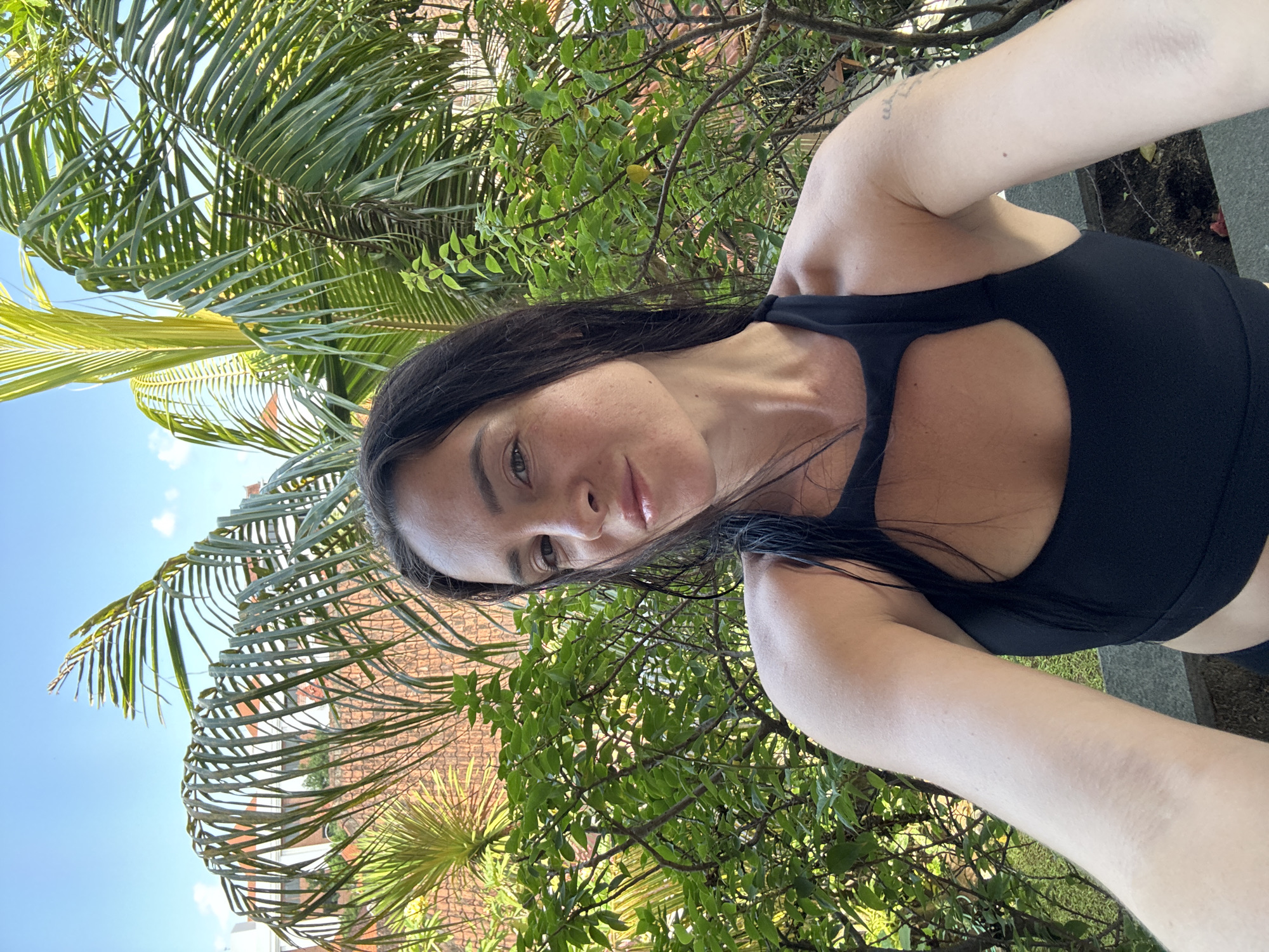 An outdoor, mid-shot, front-facing selfie shows a woman with long dark hair and fair skin wearing a black sports bra. She is smiling faintly and looking towards the camera. Behind her, lush green foliage and palm trees dominate the scene, with a glimpse of terracotta-tiled buildings and a bright blue sky peeking through. The setting appears to be a tropical environment, consistent with the area of North Kuta, Indonesia.