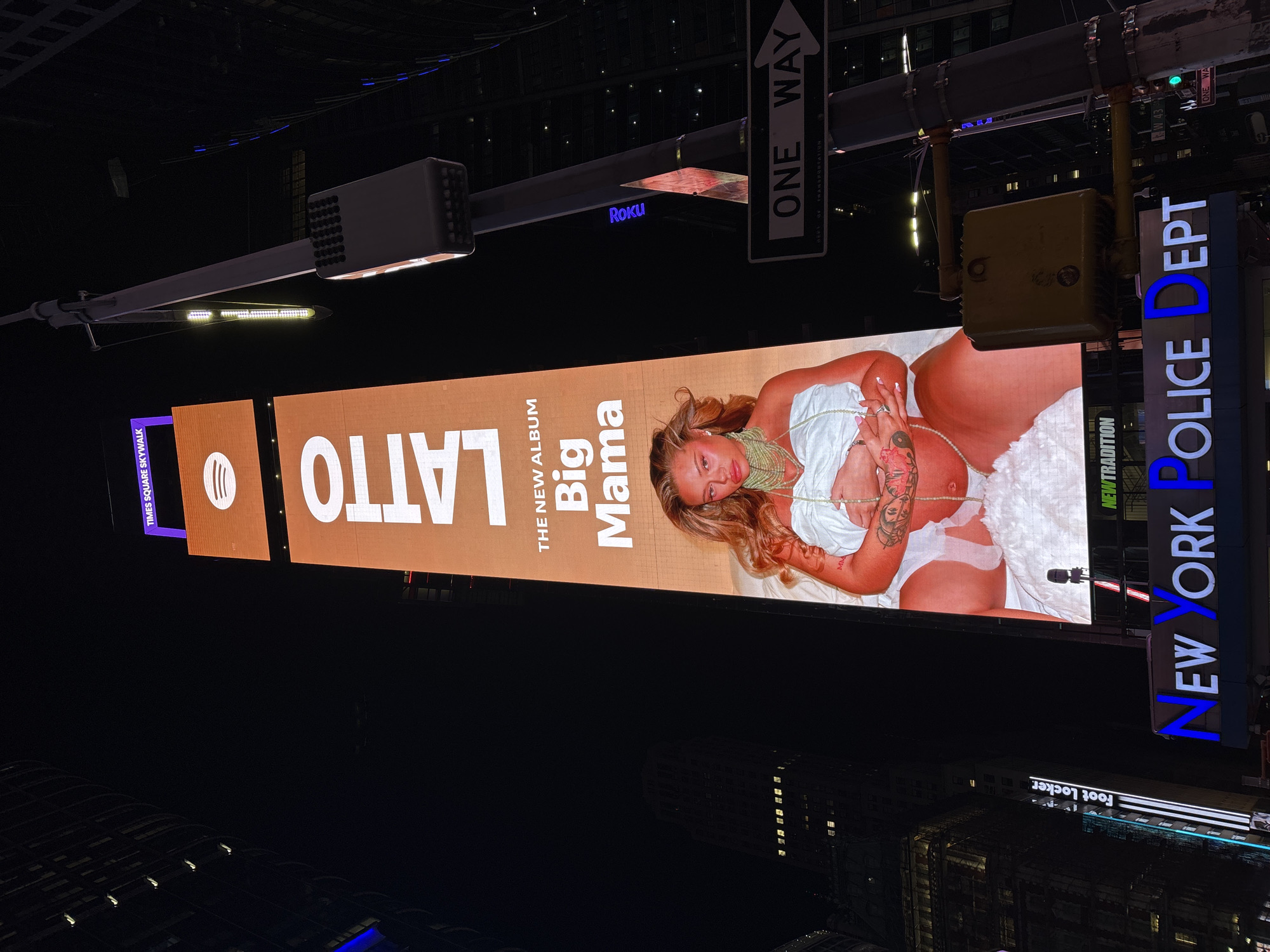 A night-time photograph captures a towering vertical LED billboard in a bustling urban environment, typical of Times Square, New York City. The image is taken from a slightly low angle, looking up at the illuminated structures against a dark sky.
The central subject is a large digital advertisement promoting "LATTO" and "THE NEW ALBUM Big Mama." The billboard features a light-skinned woman with long, wavy blonde hair, reclining or lounging. She is dressed in a white top and white undergarments, adorned with multiple green and white beaded necklaces or body chains. Tattoos are visible on her arms and torso. A Spotify logo is also displayed on the left side of the billboard.
The surroundings are filled with commercial signs and street furniture. Directly above the main billboard, a "ONE WAY" street sign is prominently displayed, alongside another sign indicating "W 43" and "ONE WAY." A dark, elongated structure with the "Roku" logo is visible. To the right of the large screen, a bright vertical sign reads "NEW YORK POLICE DEPT." Below the main advertisement, the illuminated sign for "Foot Locker" can be seen on a building in the background. Other visible text includes "TIMES SQUARE SKYWALK" and "NEW TRADITION."
The time of day is clearly night, with the sky appearing completely dark, contrasting sharply with the bright, vibrant light emanating from the numerous screens and signs. The scene conveys the energetic, commercial atmosphere of a major city at night, with advertisements dominating the visual landscape.