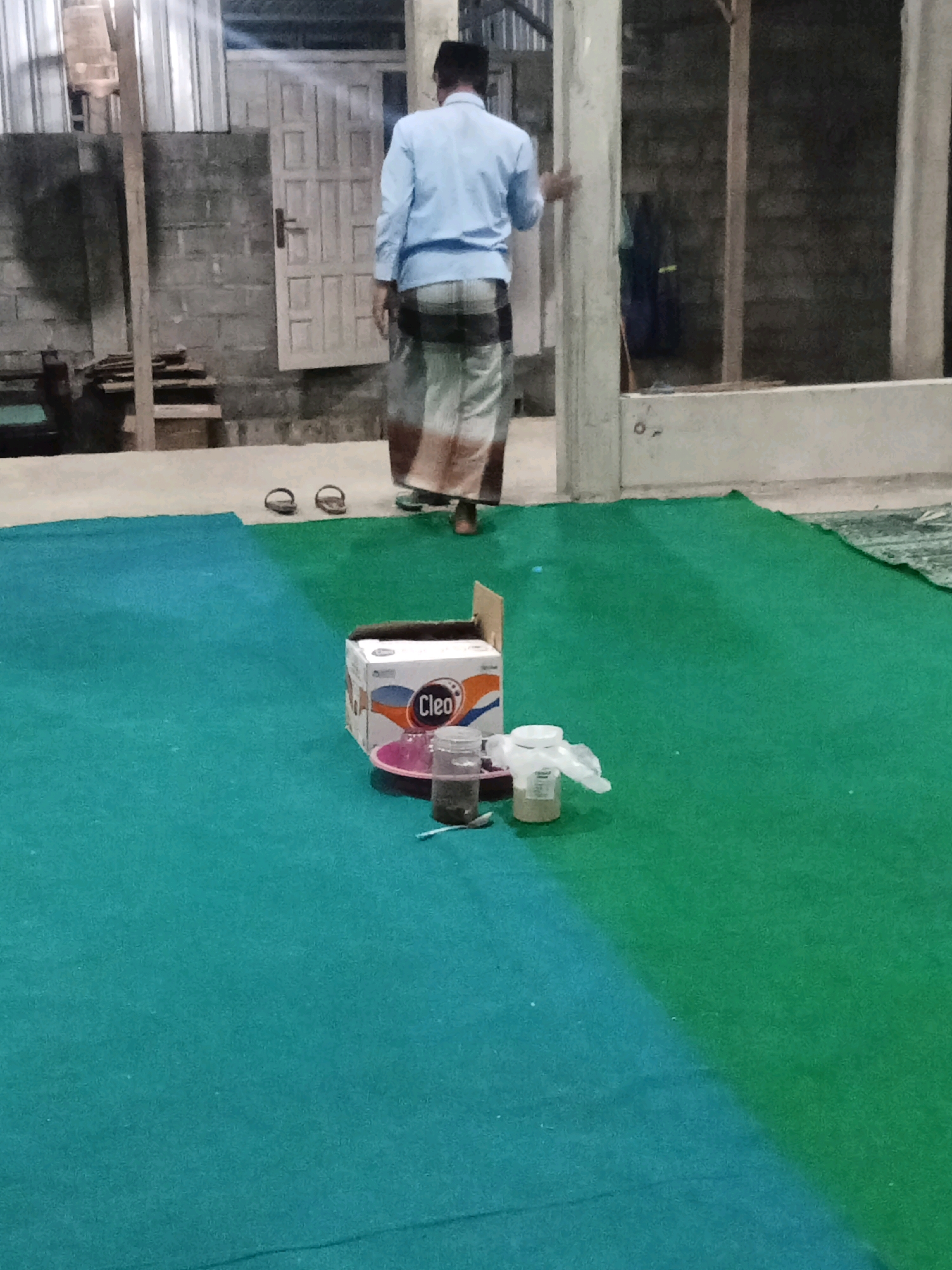 A man wearing a light blue shirt and a sarong is standing in what appears to be an outdoor or semi-outdoor location. He is facing away from the camera and appears to be walking or standing near a doorway. In the foreground, there is a large, two-toned carpet, half teal and half green. On the teal section of the carpet, there is a cardboard box labeled "Cleo," a pink plate, and two small containers. There are also a pair of dark-colored flip-flops visible on the ground behind the man. The background shows a brick wall, a wooden structure, and a metal roof. The lighting suggests it is evening or nighttime. The location is identified as Keplaksari, Indonesia.