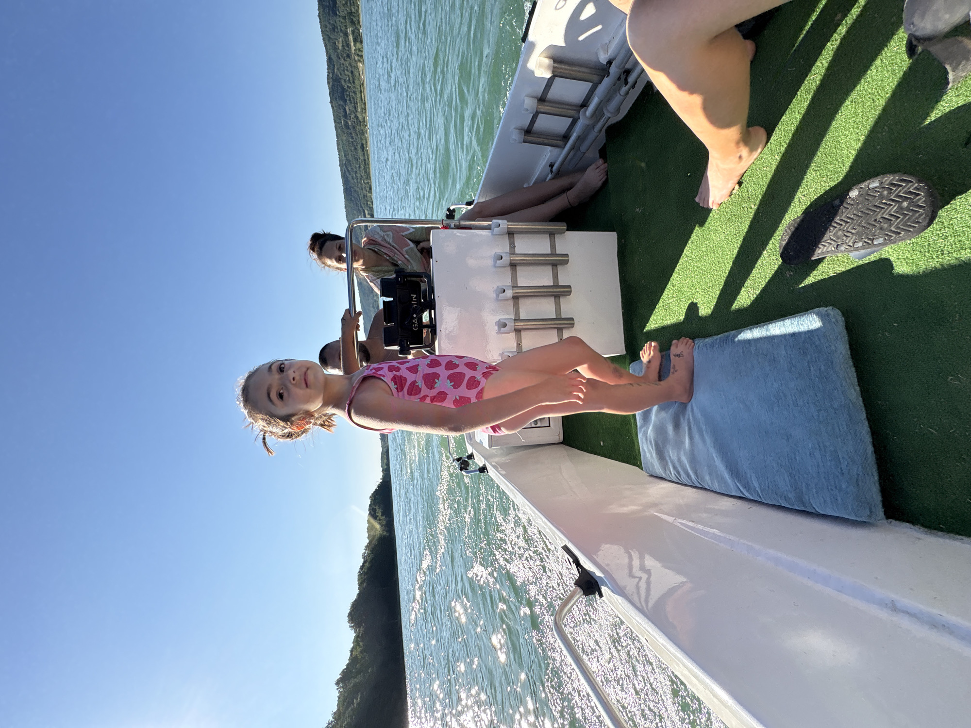 A low-angle, medium shot captures a young girl in a pink strawberry-patterned swimsuit standing on a boat's deck. She is facing forward and to the right, her gaze directed slightly upwards and away from the camera. Behind her, two adults are seated. A woman with dark hair pulled back in a bun is at the helm of the boat, wearing a light-colored, patterned garment. A man with dark skin and a shaved head is partially visible behind her. The boat is on a body of water, with the sun glinting off the surface. Green, hilly terrain lines the horizon under a clear blue sky. The boat's interior is covered with green carpeting, and several white rod holders are mounted on a console. A blue cushion is positioned on the deck near the girl. The image appears to be taken during daylight hours, with strong sunlight casting shadows. The location is identified as the city of Golyama Zhelyazna, Bulgaria.