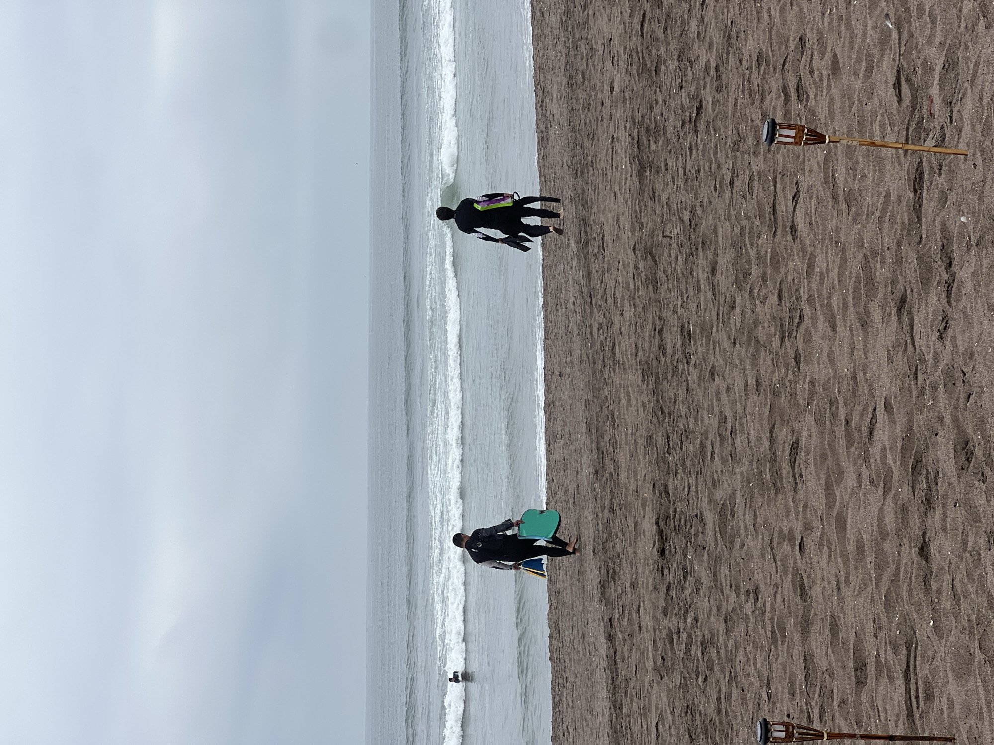 Two individuals in wetsuits walk on a sandy beach toward the ocean. The person on the left is carrying a turquoise bodyboard and a blue and yellow swim fin. The person on the right is carrying black swim fins and a green bodyboard. In the distance, another individual is in the water. The sky is overcast, and the ocean has moderate waves. Two tiki torches are planted in the sand in the foreground. The scene appears to be at the urban coast of Arica, Chile, likely near the Playa El Laucho or Playa La Lisera, given the presence of these amenities.