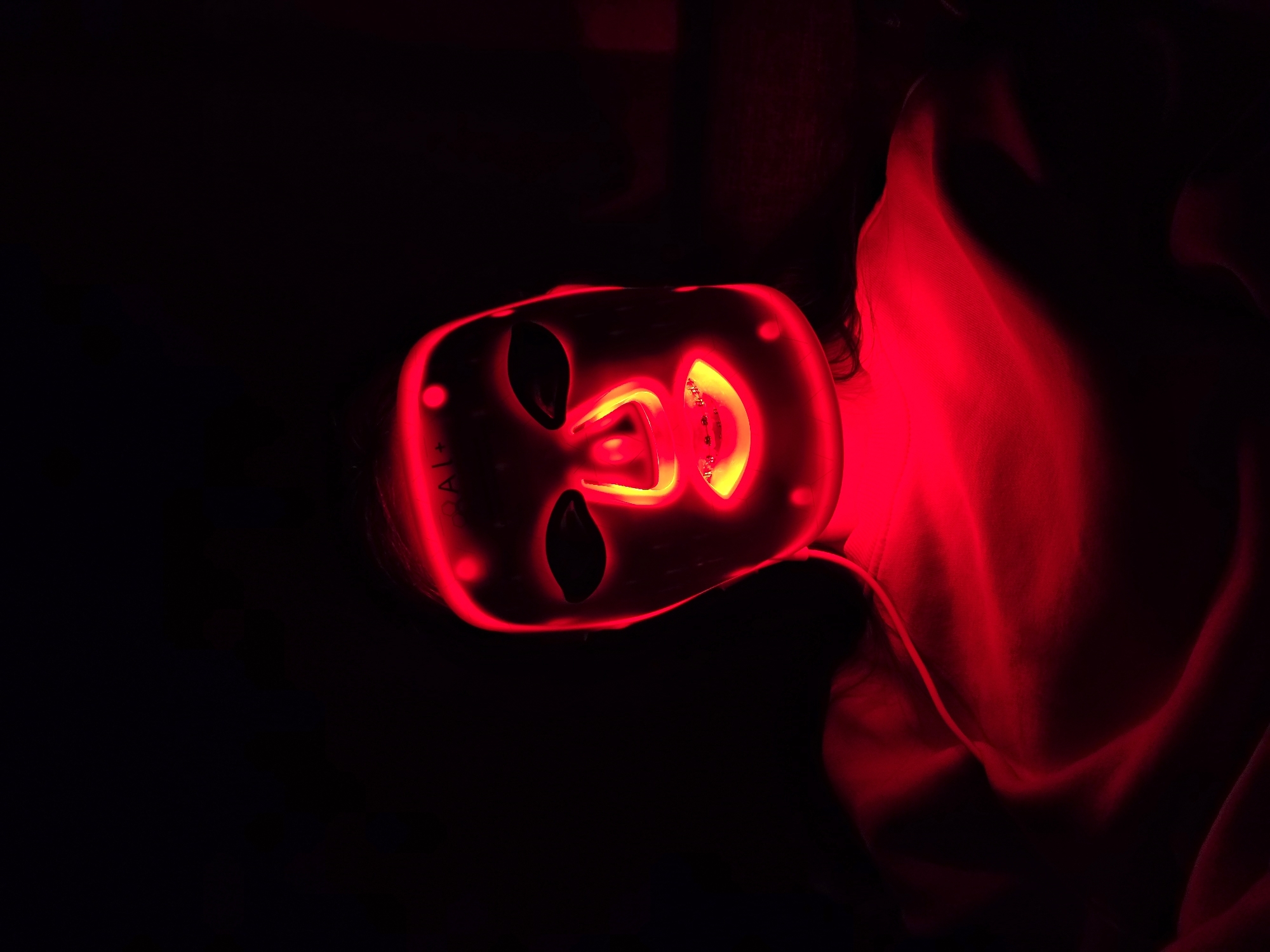 A person is wearing a red LED light therapy mask. The room is dark. A red article of clothing can be seen on the person.