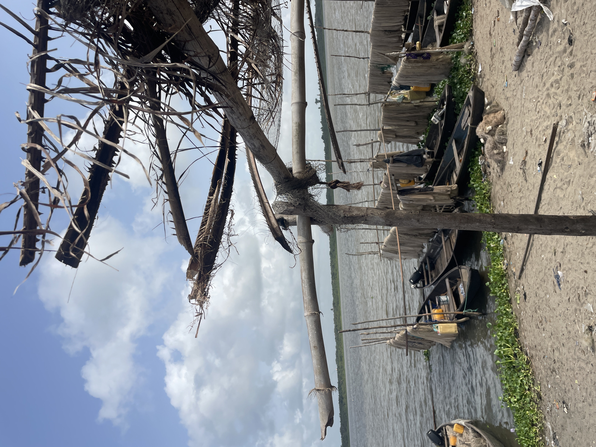 The image shows a waterfront scene in an area characterized by simple structures and boats. In the foreground, wooden beams and branches form part of a structure, possibly a shelter or dock. Beyond the structure, the water surface is visible, reflecting the sky. Along the shore, a series of boats are docked, some partially covered by nets or cloths. The shore area is sandy and appears to be littered with debris. The background features a distant shoreline with vegetation. The sky is visible through gaps in the foreground structure, showing blue with scattered clouds. The image's orientation is vertical.