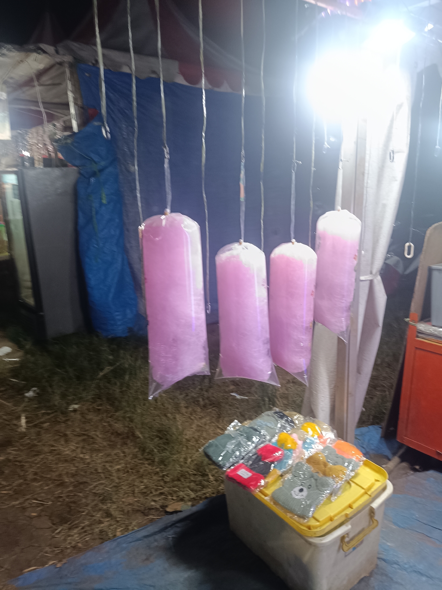 This media file depicts an outdoor stall likely at a night market or fair in Depok, Indonesia. The primary subjects are several large, clear plastic bags filled with fluffy pink cotton candy, suspended from strings. The cotton candy appears to be in various stages of being formed, with some bags fully packed and others less so. To the right of the cotton candy, there is a yellow plastic container overflowing with small, individually packaged items, likely souvenirs or novelty goods. These items are wrapped in clear plastic and include what appear to be small hats or beanies with embroidered designs, such as a grey bear face. Other brightly colored items are also visible within the container.
The setting is dimly lit, with a bright, harsh light source, possibly a floodlight, dominating the upper right portion of the image, creating lens flare and washing out some details. This suggests the scene is illuminated for evening or nighttime activity. The ground appears to be covered in dry grass or hay, and there are tarps or fabric coverings in the background, indicating a temporary stall structure. Behind the cotton candy, large blue and white tarps are visible, possibly serving as backdrops or shelter.
The overall atmosphere seems to be that of a bustling market. While no people are clearly visible in the frame, the presence of merchandise and the stall setup implies commercial activity. The time of day is clearly night or late evening due to the artificial lighting. There are no overt emotional cues conveyed, as the focus is on the products and the stall itself. No visible text is discernible in the image. The context of Depok, Indonesia, further situates this as a scene from a local Indonesian market.