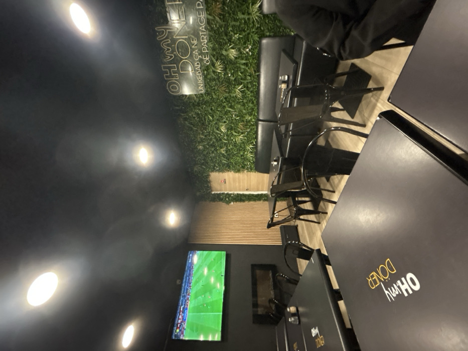 The scene depicts a modern restaurant interior. Black tables with branding are prominent, accompanied by black chairs. A long black banquette lines a wall adorned with artificial green foliage. A television displaying a soccer match is mounted on the opposite wall, which features a wood-paneled section. Overhead lighting fixtures illuminate the space.