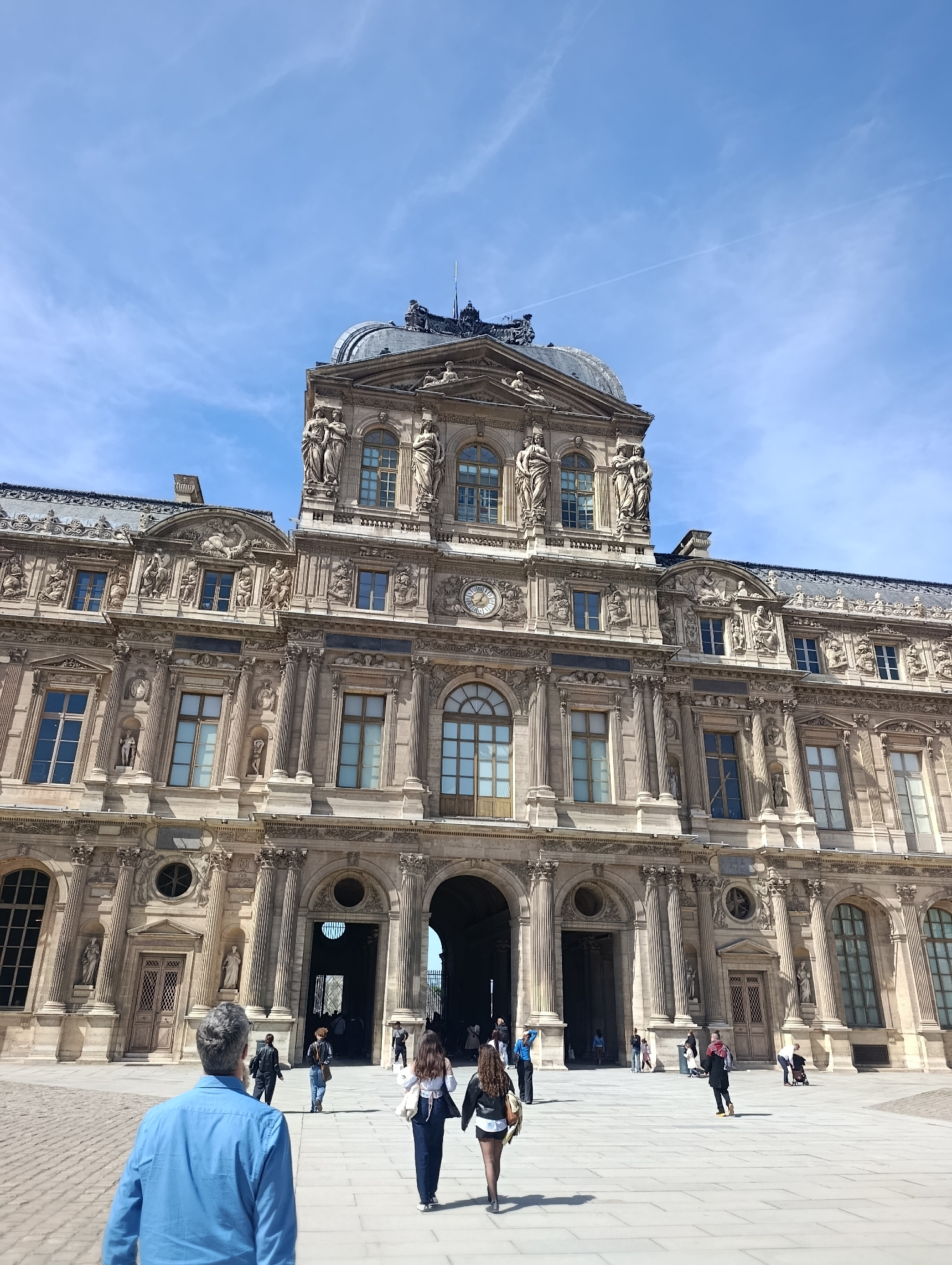 The image shows the Louvre Palace in Paris, France. The building is ornate, with a dome on top, statues, and many windows. People are walking in front of the building. The sky is blue with some clouds. The foreground shows a courtyard paved with stone slabs.
