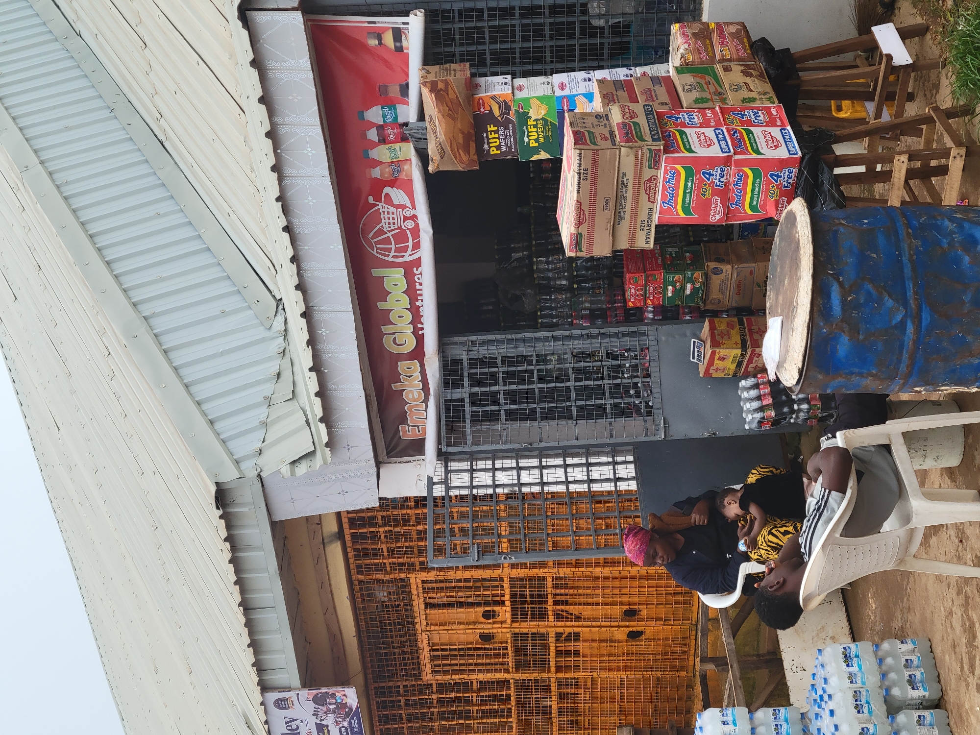 The image shows a storefront in Bokkos, Nigeria, with a red banner that reads "Emeka Global Ventures". In front of the store, there are stacks of water bottles and boxes of Indomie noodles. A woman wearing a pink headscarf is sitting in a white plastic chair, holding a baby. A young man is lying back in another white plastic chair, his eyes closed. The ground is dirt and gravel, and there are several metal barrels visible. The overall scene suggests a small business operating in an outdoor setting.