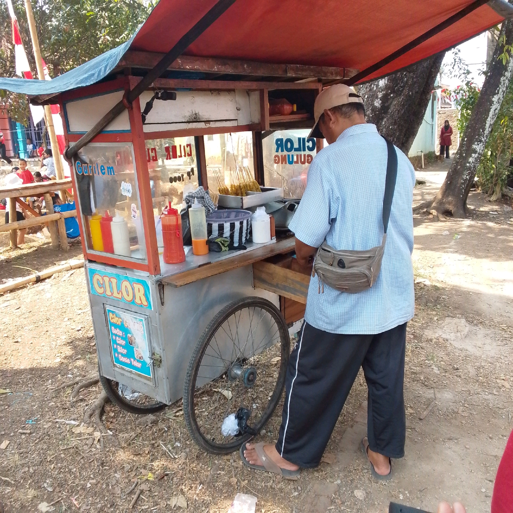 A street vendor is preparing food at a cart labeled "Cilor" in Cilaku, Indonesia. The cart is made of metal and wood, with a red canopy. It is filled with various food items, including skewers of food and bottles of sauce. The vendor, wearing a blue plaid shirt and black pants, is seen from the back as he works at the cart. In the background, there are other people and trees, suggesting an outdoor setting. The ground is covered in dirt and fallen leaves.