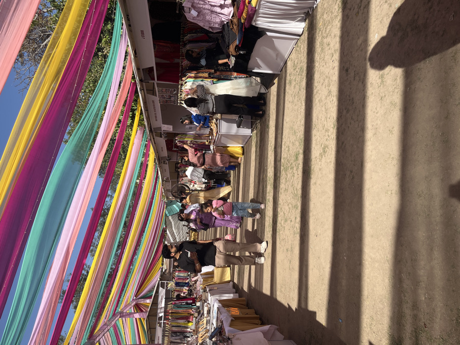 The scene is set in a marketplace in Sector 17, Chandigarh, India. The image captures a street lined with market stalls adorned with colorful fabric strips. The ground is a mix of dirt and gravel, with visible shadows cast by a canopy of colorful fabric strips. There are several people walking along the street, some browsing the stalls. A young girl in a pink dress and a young boy in a black shirt are walking hand-in-hand on the left side of the image. The market stalls are stocked with various products, including clothing, jewelry, and other items. The air is clear and bright, with a blue sky and patches of cloud. A large shadow can be seen in the center of the street.