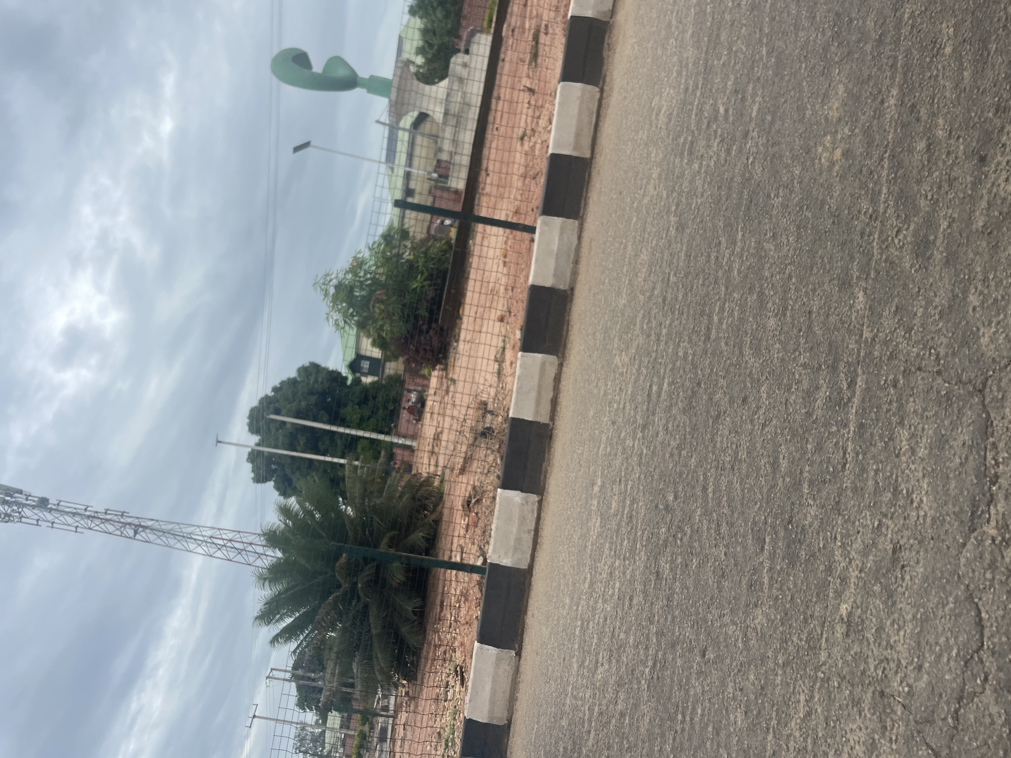 The image captures a street view in Jos, Nigeria, with a focus on a roadside roundabout. In the foreground, a cracked asphalt road leads the eye towards the roundabout, marked by a black and white striped concrete curb. Beyond the curb, a fenced-off area with sandy soil is visible, populated by several trees, including a prominent palm tree. A tall telecommunications tower stands in the background, its lattice structure silhouetted against a cloudy sky. To the right, a distinctive green sculpture, possibly representing a telephone receiver, is situated atop a circular concrete base within the roundabout. A building with a green roof and an upper floor is partially visible behind the trees. The scene appears to be set during the daytime, under an overcast sky, suggesting a potentially humid climate. No people are clearly identifiable in the image, although a blurred figure on a motorcycle is faintly visible in the far background to the right.